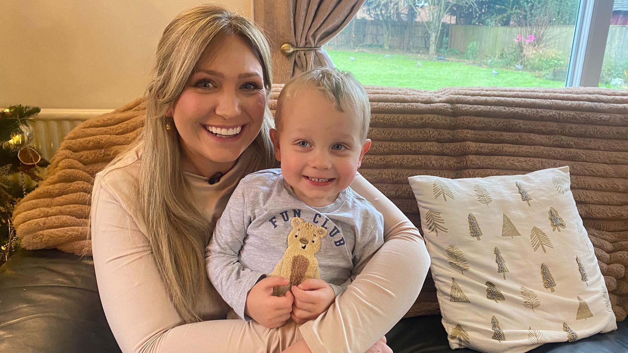 A picture of a blonde woman wearing a light pink top sat on a brown sofa, cuddling her two-year-old son who has blonde hair and blue eyes, smiling at the camera.