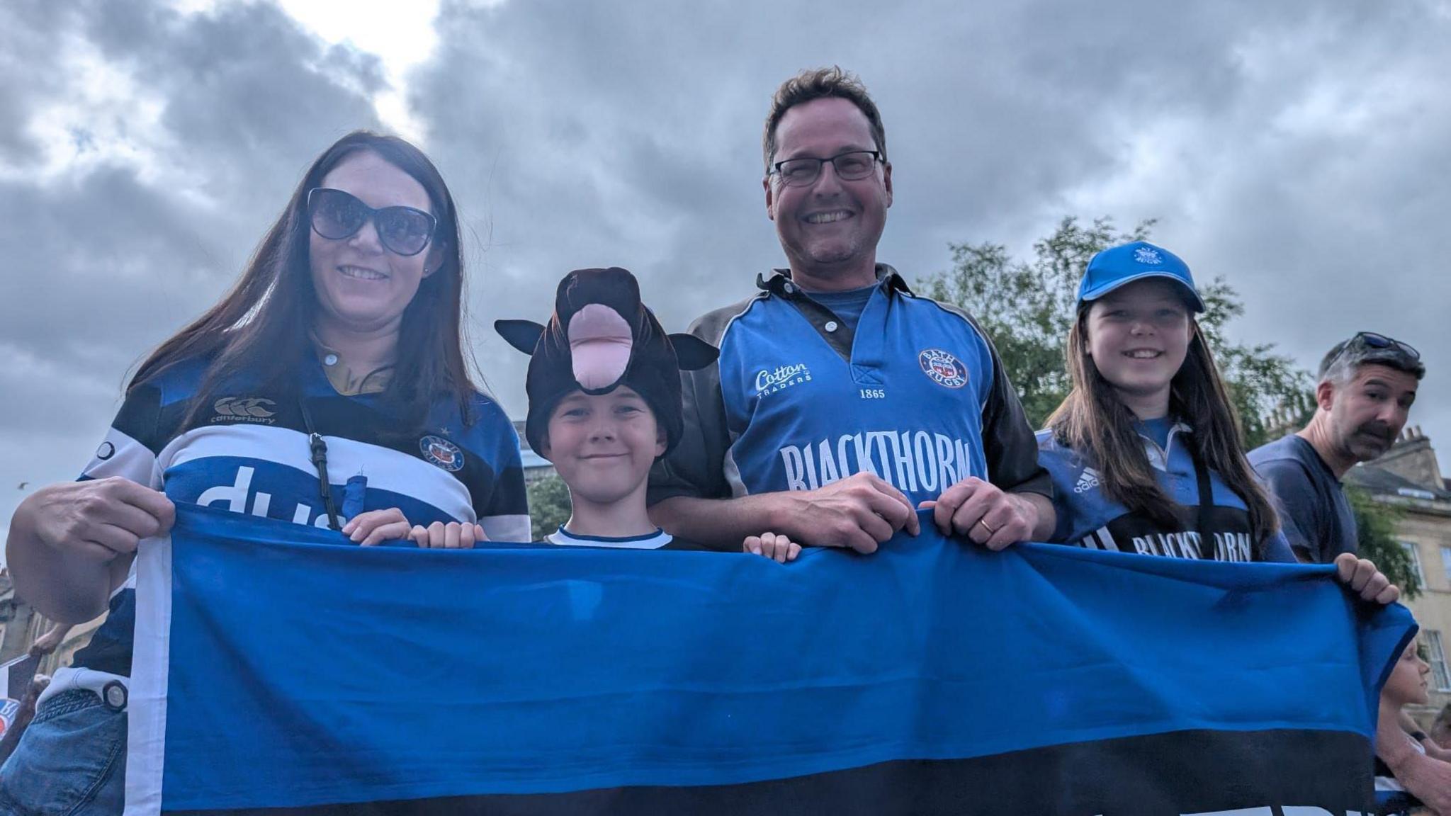 A family of four holding the top of a Bath flag and smiling at the camera