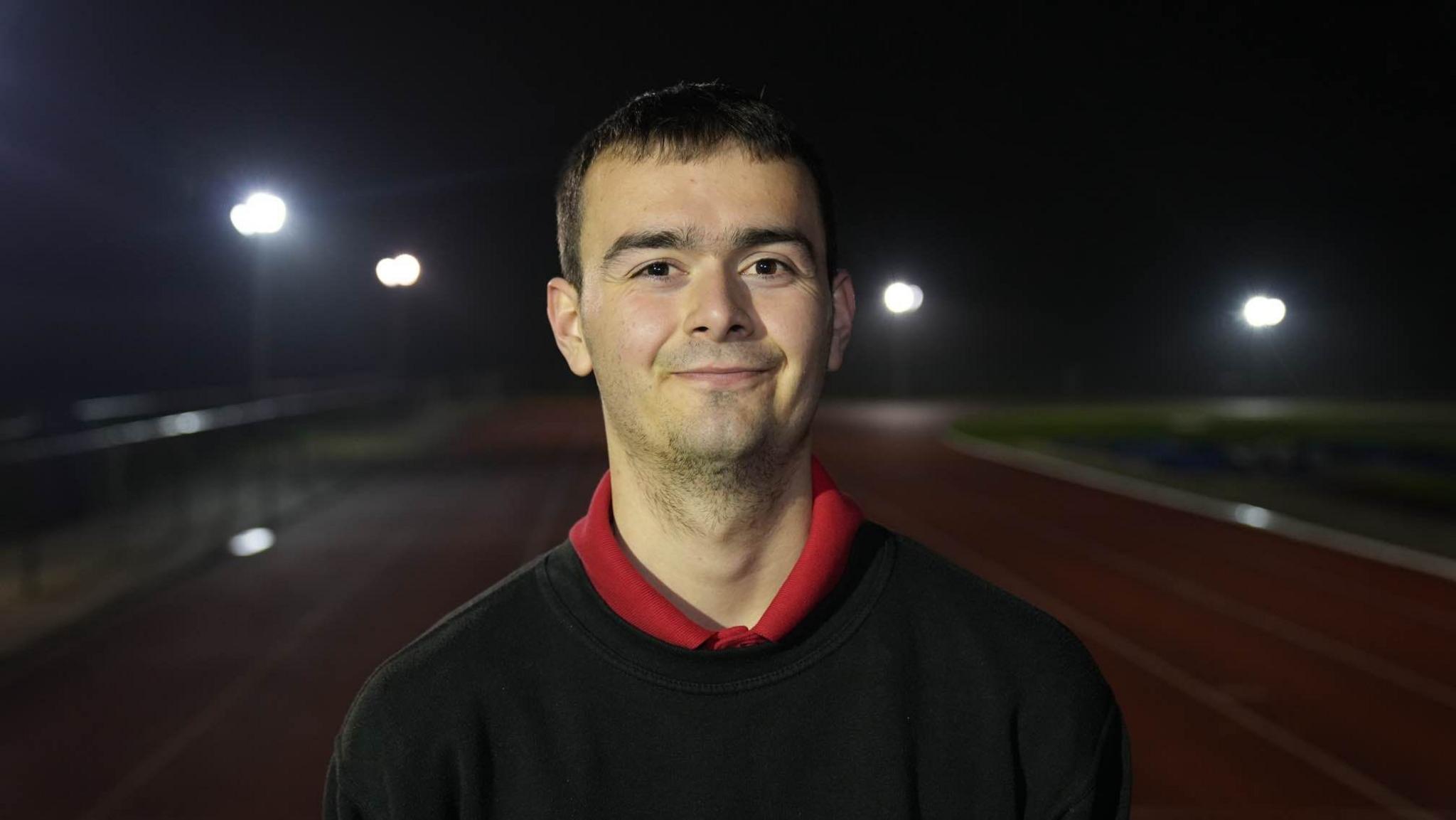 Rhys Ford smiles at the camera while standing on the athletics club. He has short dark hair and some stubble. He is wearing a black jumper with a red polo top on underneath.