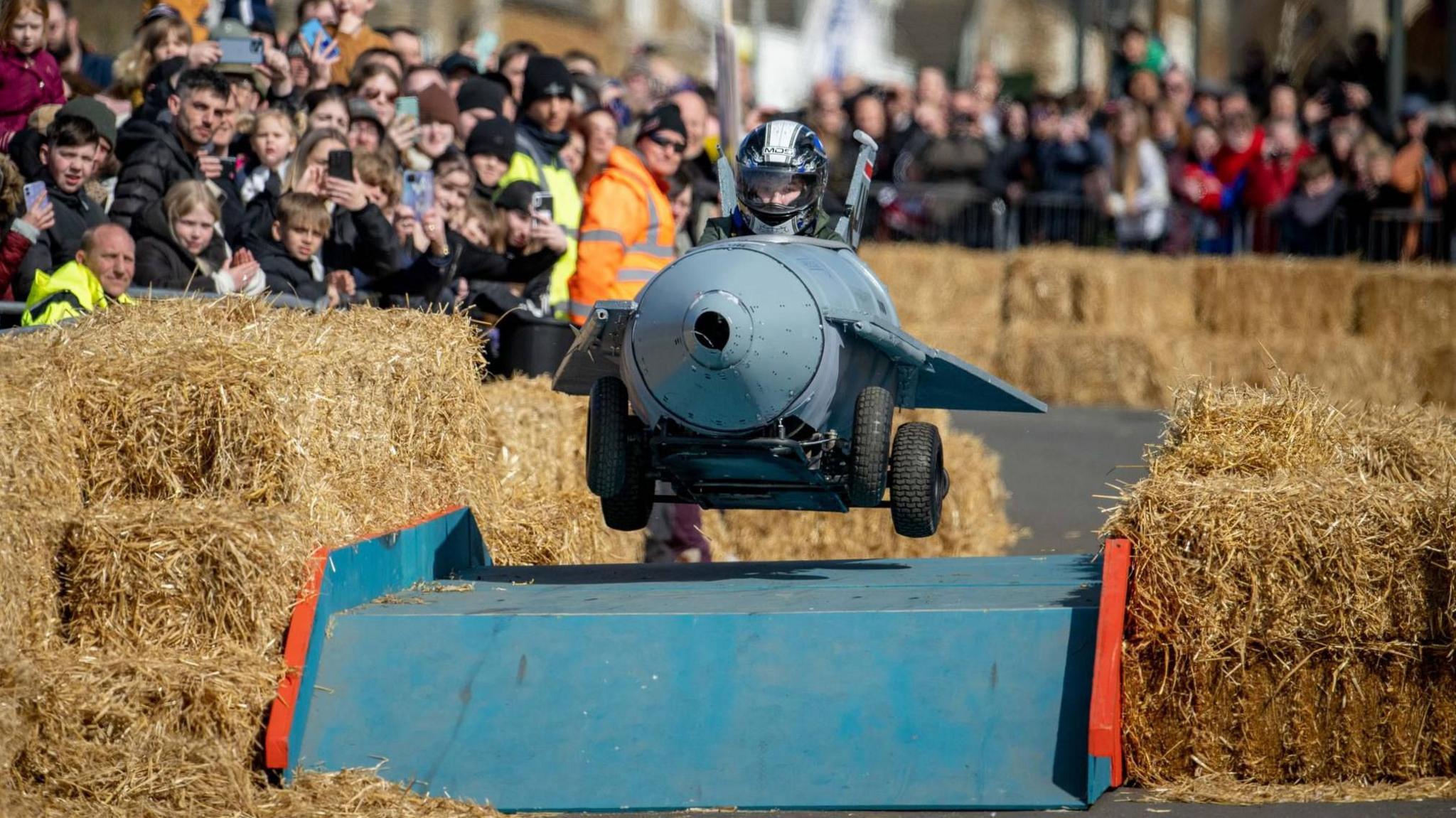 A kart that looks like a jet jumping over a ramp with hay bales either side and dozens of spectators looking on