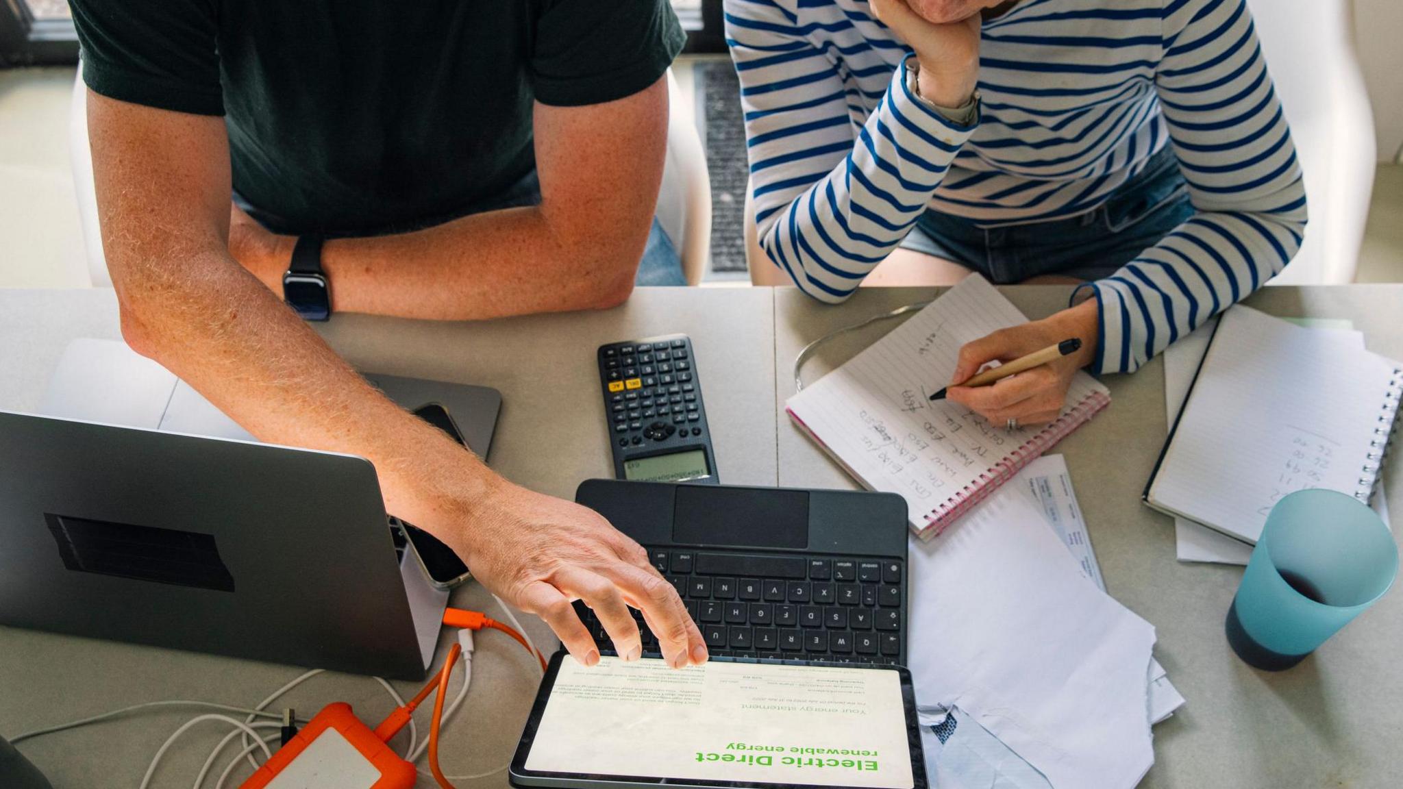 A couple sit at a large table in front of a laptop with a calculator and notebooks. They look to be going over a budgets and finances. The man is dressed in a dark t-shirt while the woman has a long-sleeved striped top.