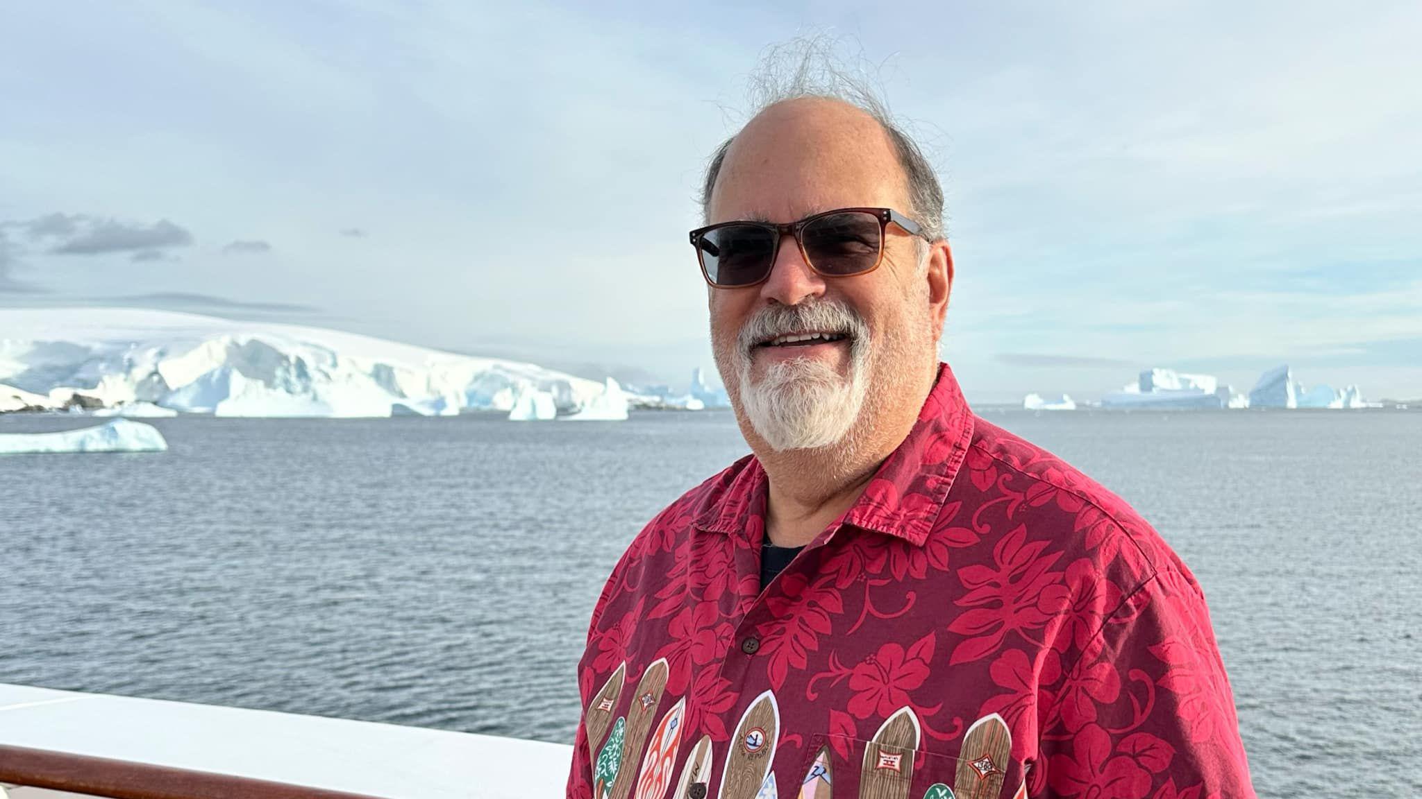 Man wearing bright red shirt with surf boards and flowers on it, wearing sunglasses. Standing in front of icebergs and the sea.