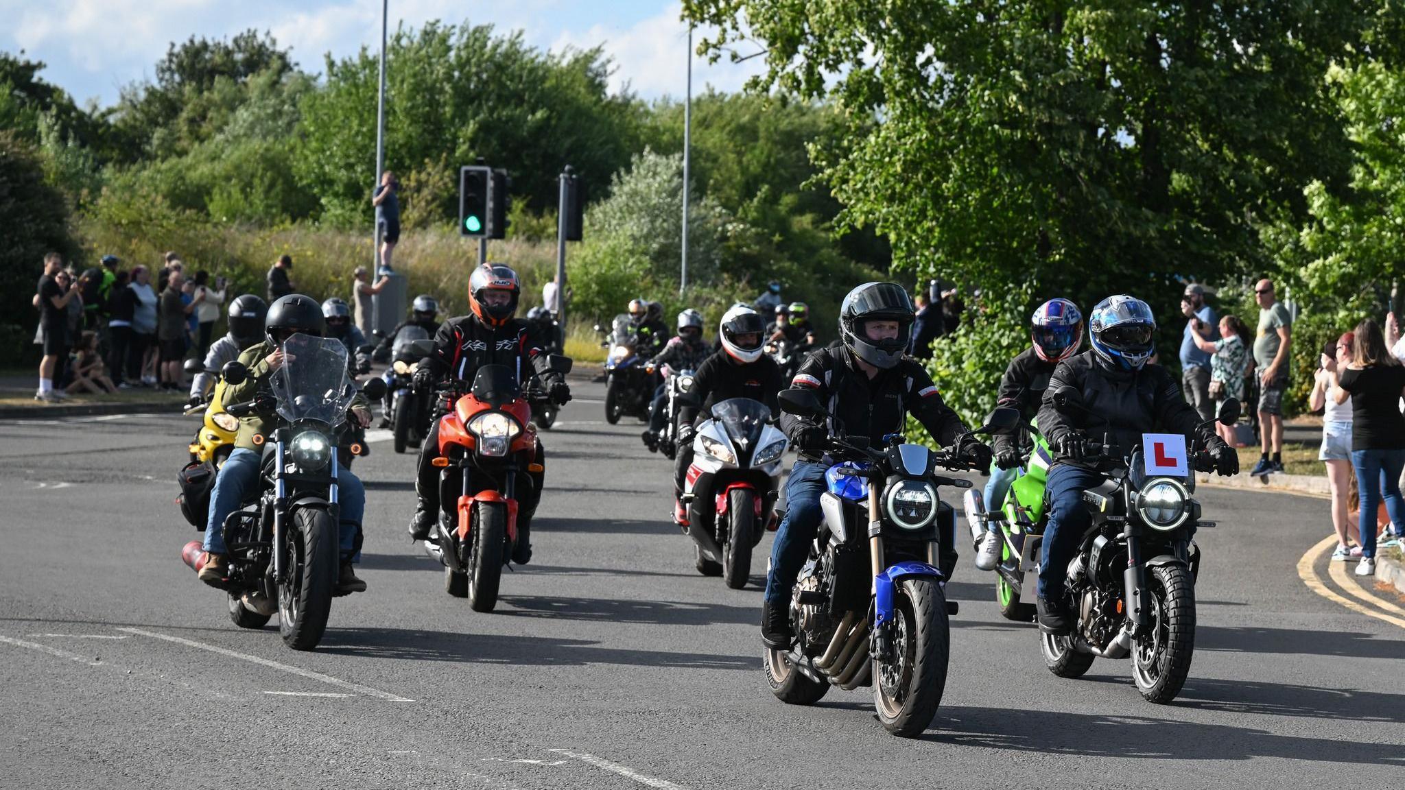A large convoy of motorcyclists driving down the road while spectators look on from the pavements.