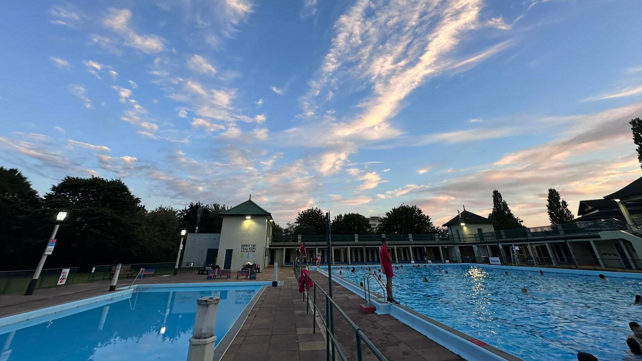 An image of the lido at sunset, it's summer and there are lots of people in the pool, the sky is red in the background.