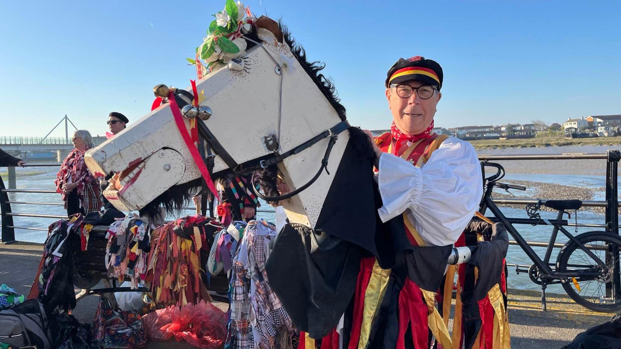 A man looks at the camera and smiles. He is wearing a Morris outfit including red and yellow silk ribbons and is holding a large wooden horse head also adorned with bells and flowers.