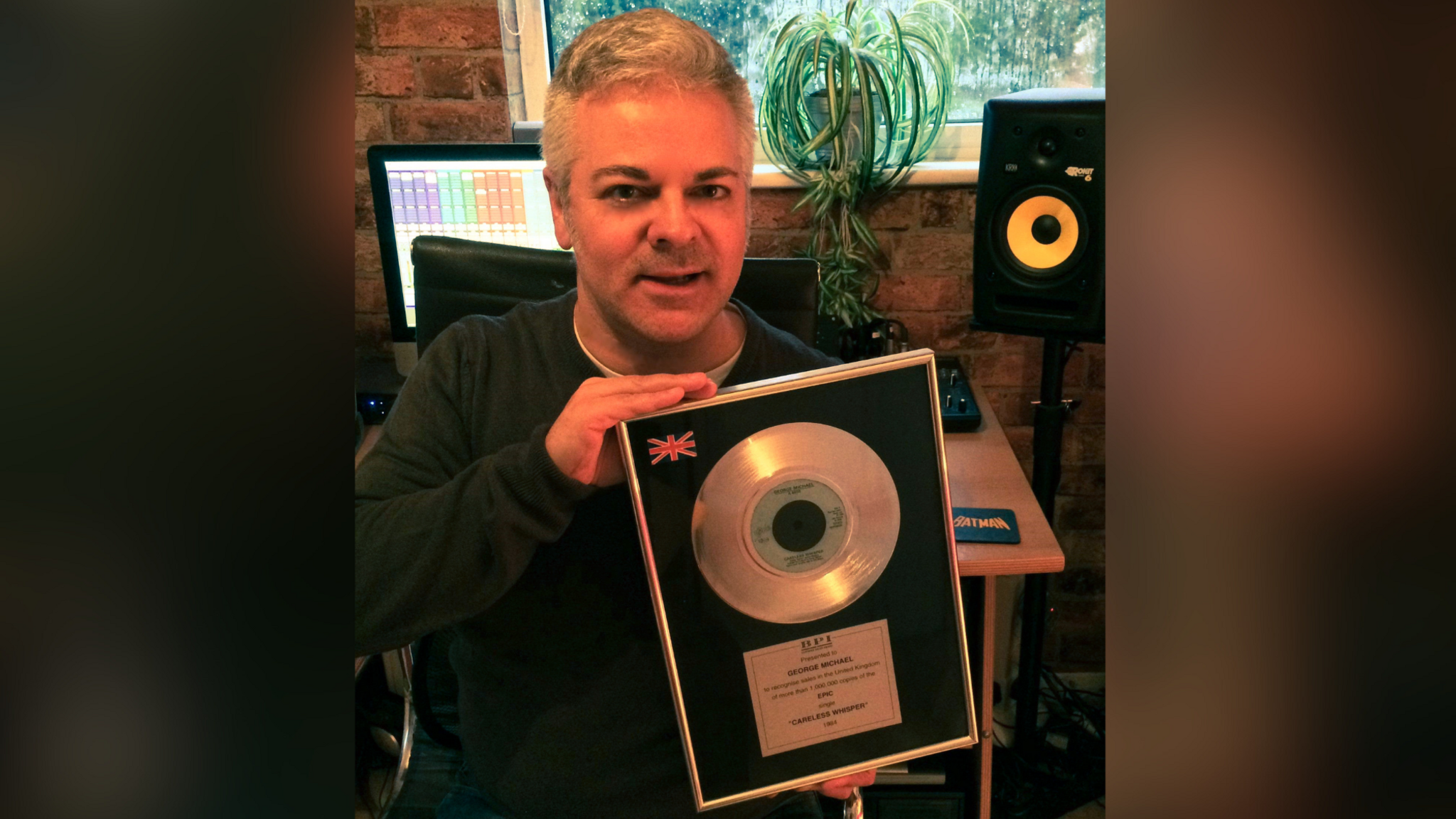 A picture of music producer Paul Mex at his Brighton studio holding the original demo of the Wham hit Careless Whisper. Paul is sitting in front of a computer and speaker, and is holding the demo. Paul has graying hair and is wearing a green jumper.