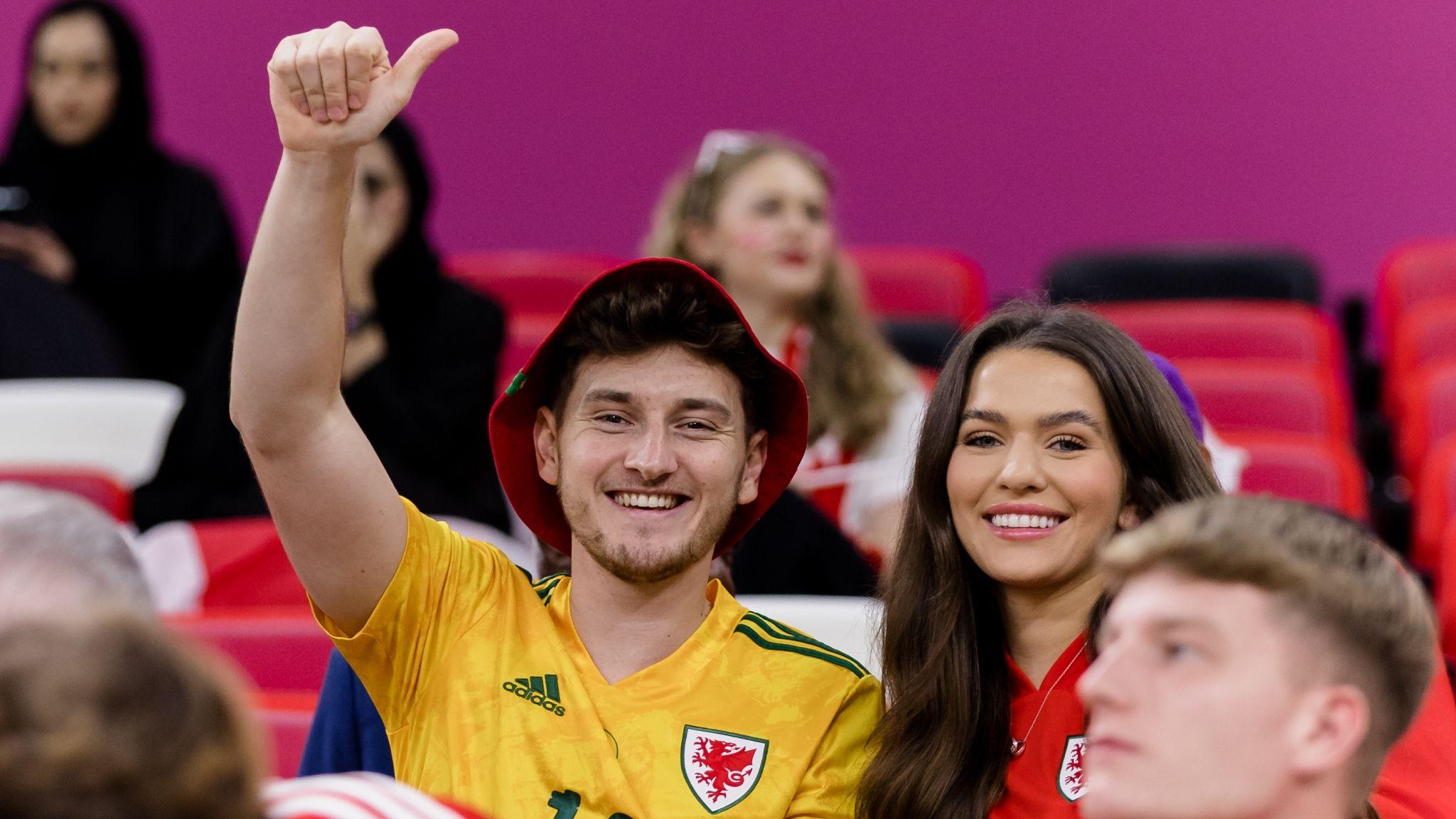 David Brooks wearing a bucket hat and a Wales shirt in the stand at Qatar 2022 raises his arm with a thumbs up
