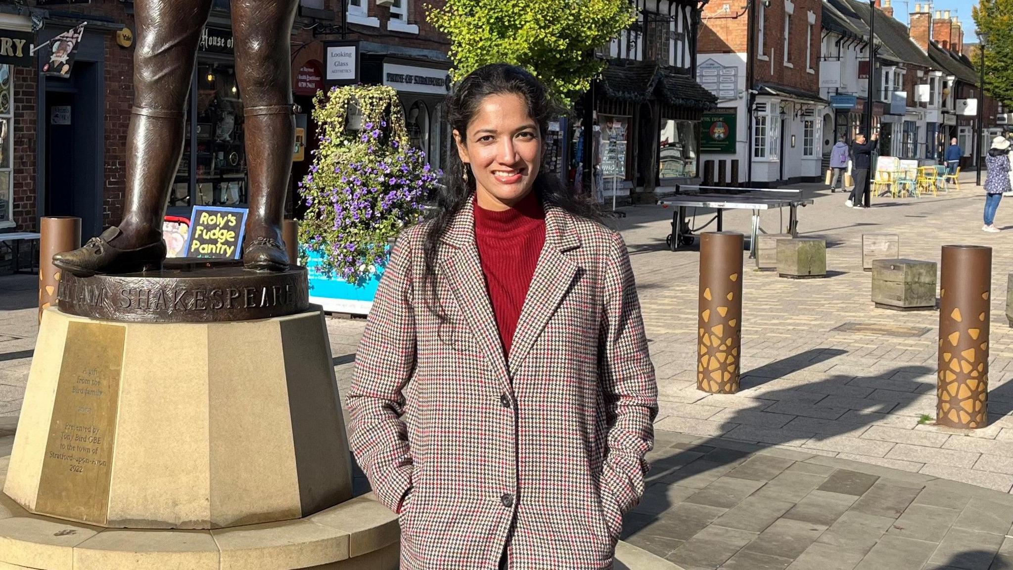 Lakshmi Balakrishnan stood with a brown coat over a red jumper. Stratford-Upon-Avon high street is behind her, including the base of a statue of William Shakespeare.