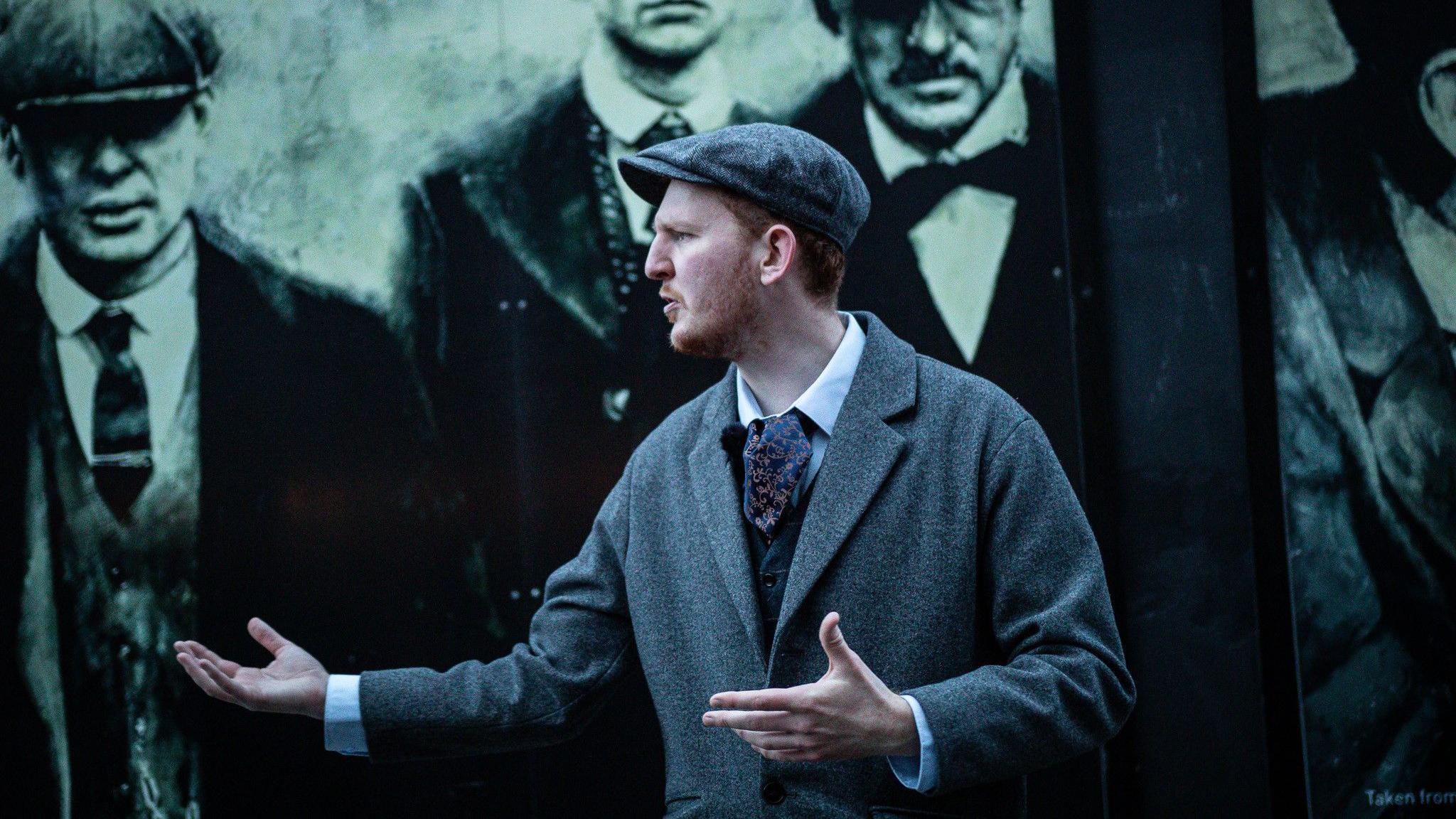 A man wearing a grey flat cap and coat stands with his arms outstretched. A peaky blinders mural can be seen behind him.