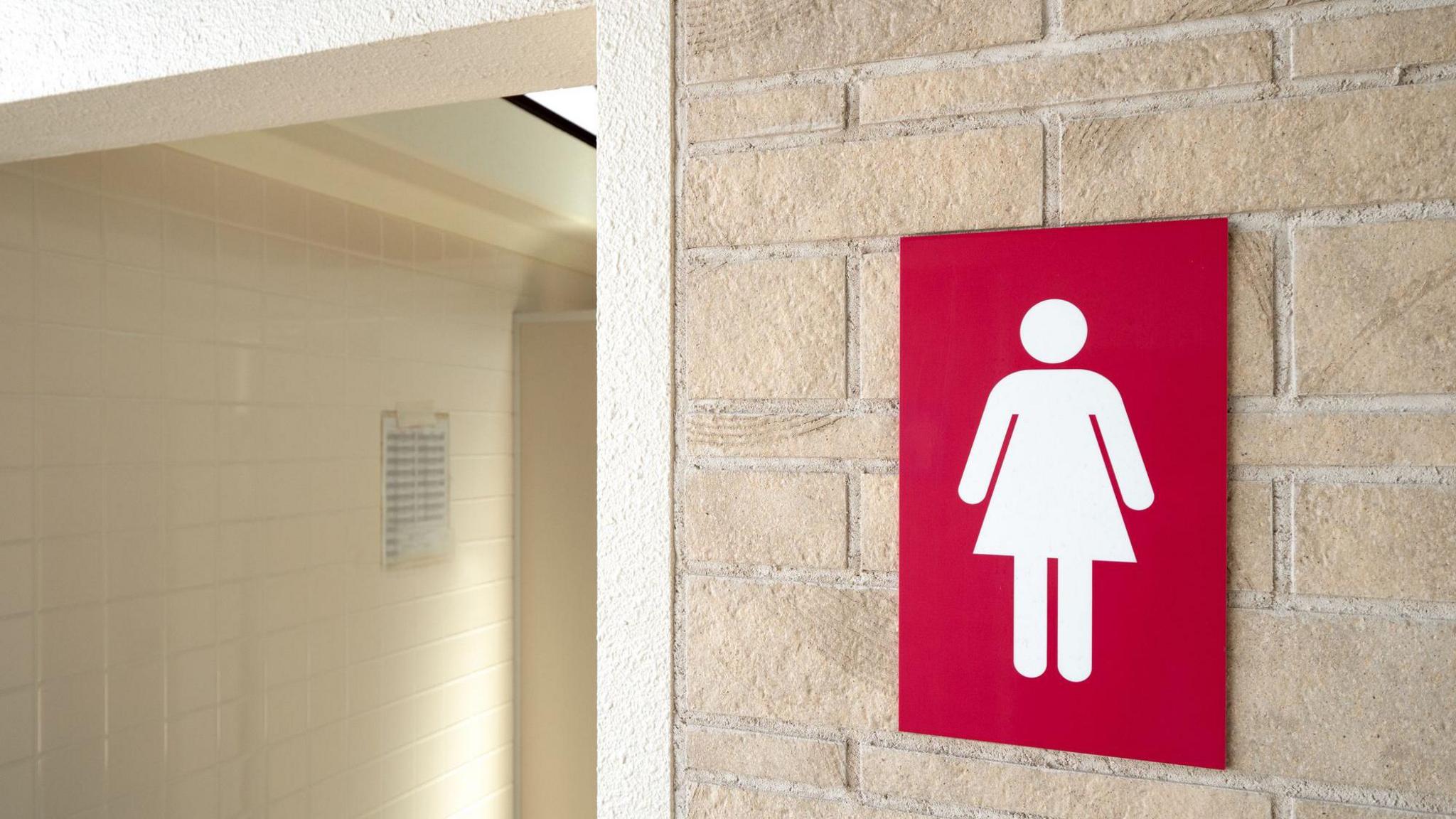A generic photo of a ladies' toilet sign on a wall outside a toilet. The sign has a white lady symbol on a red background. The doorway has pale cream tiles.