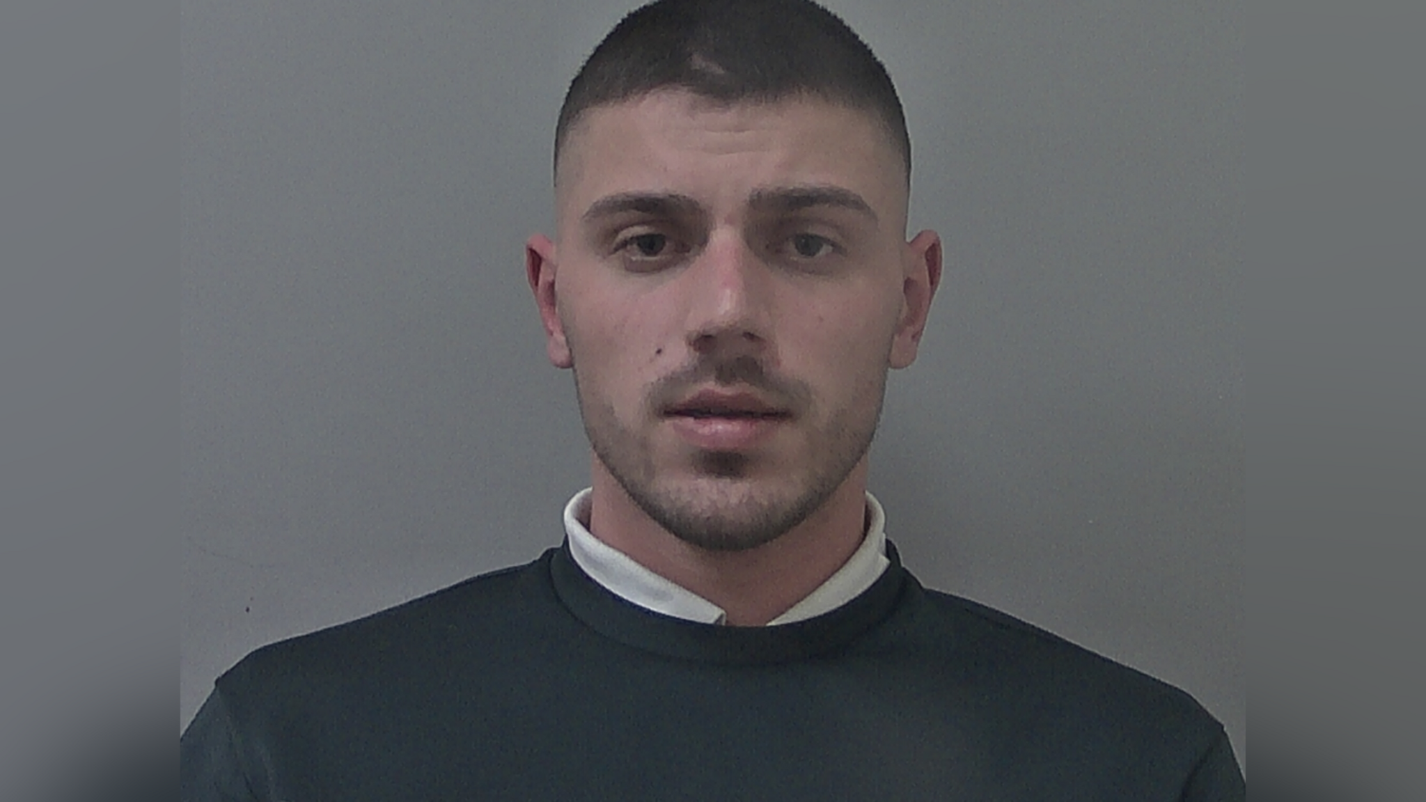 Man with dark short cropped hair and stubble. He is wearing a white shirt underneath a dark jumper.