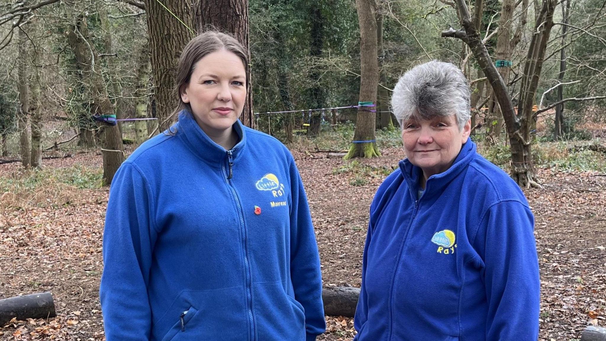 Mairead Cheeseman, left, and Pauline Cheeseman. Both are wearing blue fleeces with a yellow and blue logo of a blue cloud and yellow sun. They are standing in front of a forest clearing with trees with a zipwire tied between them.