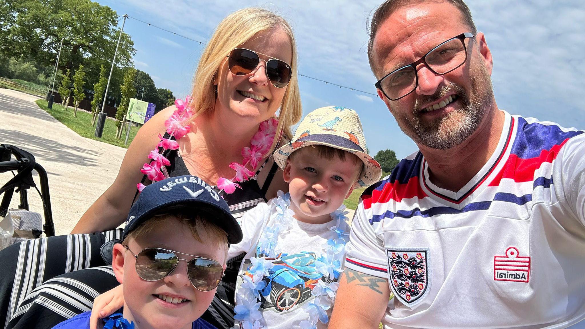 A family photo showing a man and woman smiling with two younger children on a sunny. They are all wearing sunglasses and t-shirts. The woman and the younger boy wear flower hula garland's around their neck. 