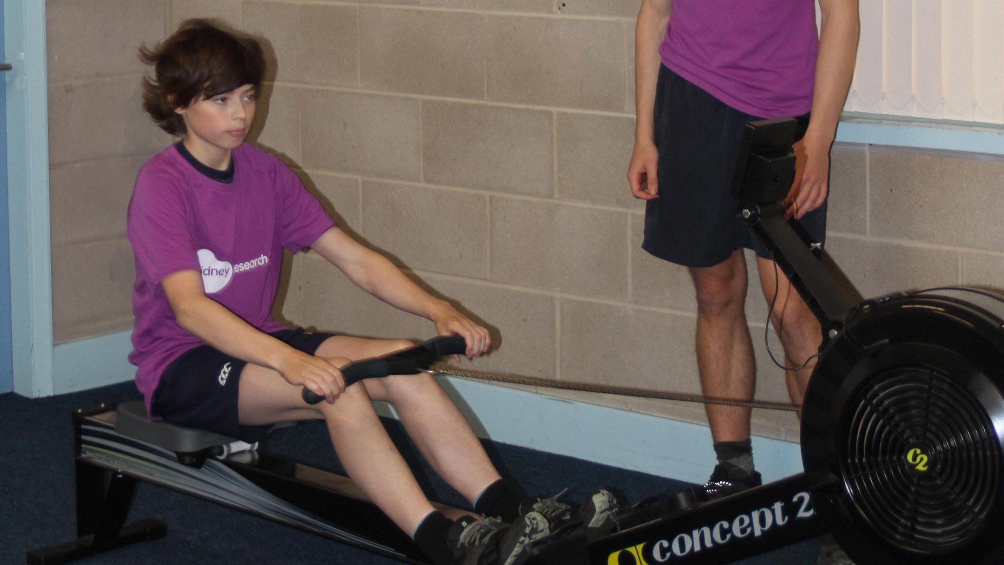 A teenage boy with brown hair and wearing a purple t-shirt and black shorts pulling at a rowing machine.