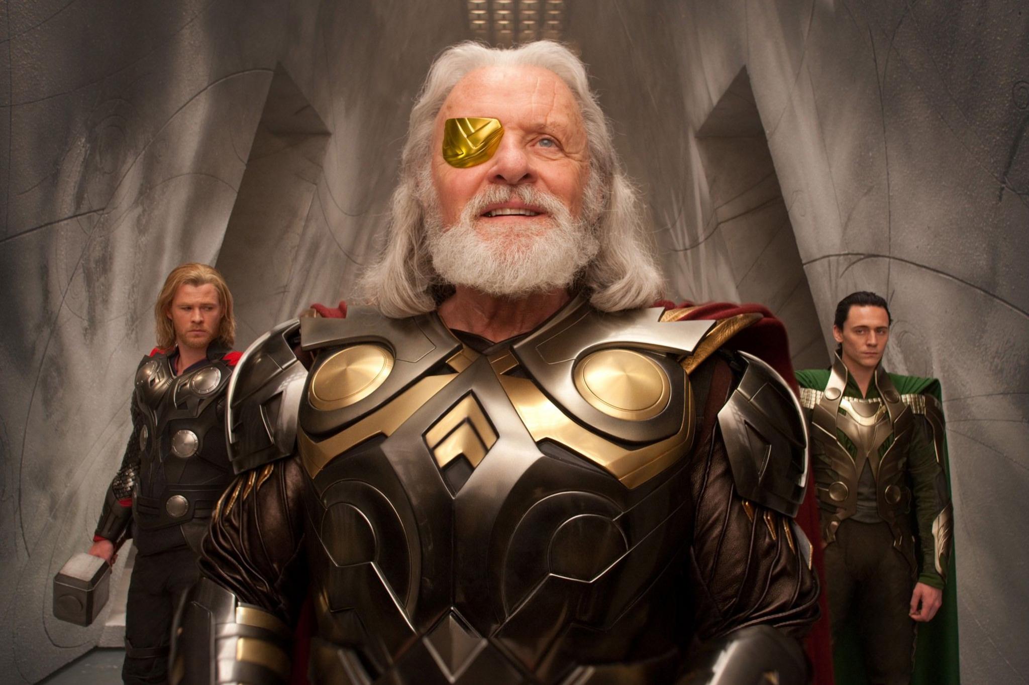 (L-R) Chris Hemsworth in the background in brown and gold armour, blond hair as Thor, Anthony Hopkins as Odin in the centre, with a gold patch on his right eye, shoulder length white hair and beard, and Tom Hiddleston as Loki, on the right in the back, black hair, gold and green armour in Thor