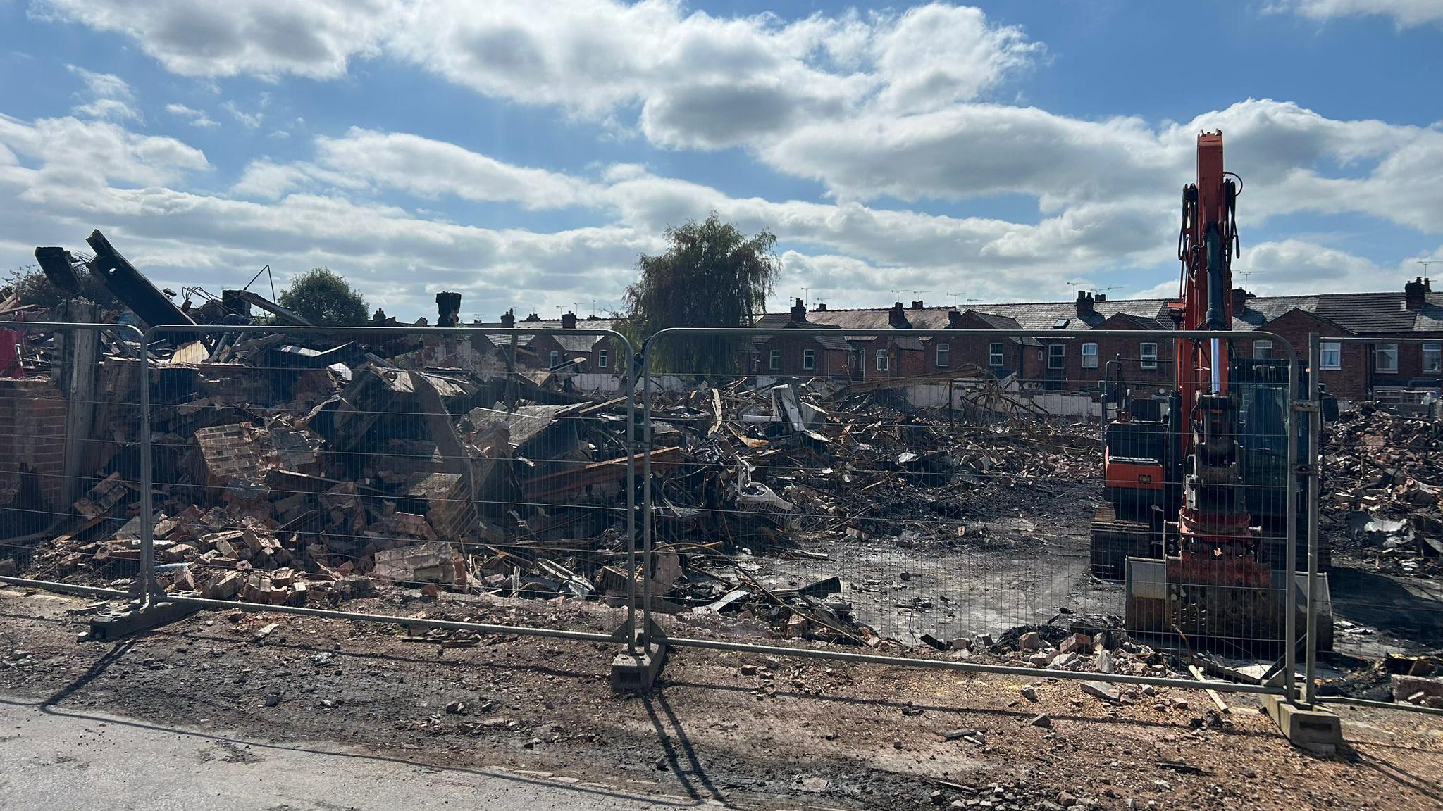 A site of a demolished building behind metal fencing. Huge piles of rubble are scattered across the site and on the right, a yellow digger is parked. Behind the site is a row of terraced houses.