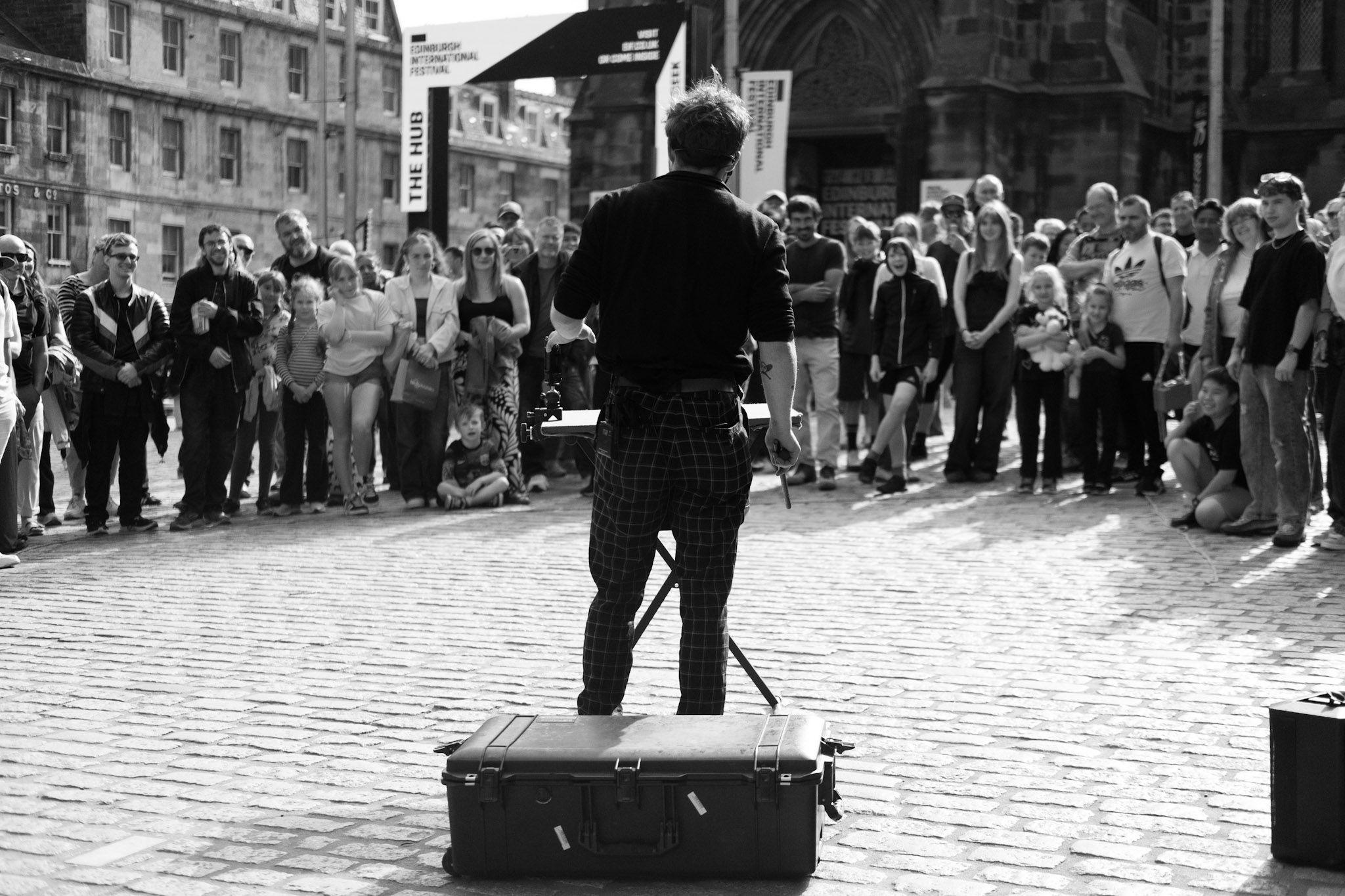 A black and white image taken from behind a street artist as he performs to a circle of people on a cobbled square in Edinburgh. A suitcase sits behind him on the ground, a table in front where he seems to be performing a trick of some kind