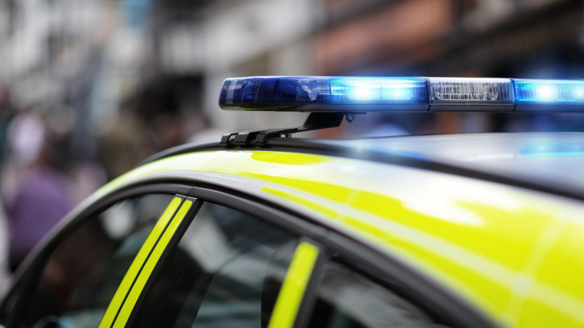 A close-up of a police car with a bright yellow and blue design. The flashing blue emergency lights are on, mounted on the roof. The background is blurred, showing a city street with people and buildings.