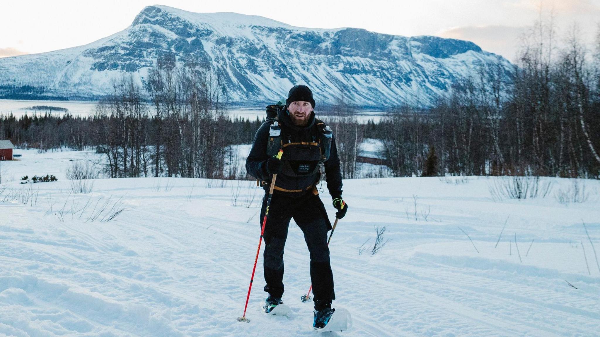 A picture of Marc Rhodes with walking poles and dressed up with warm clothes walking through snow and a large mountain behind him.