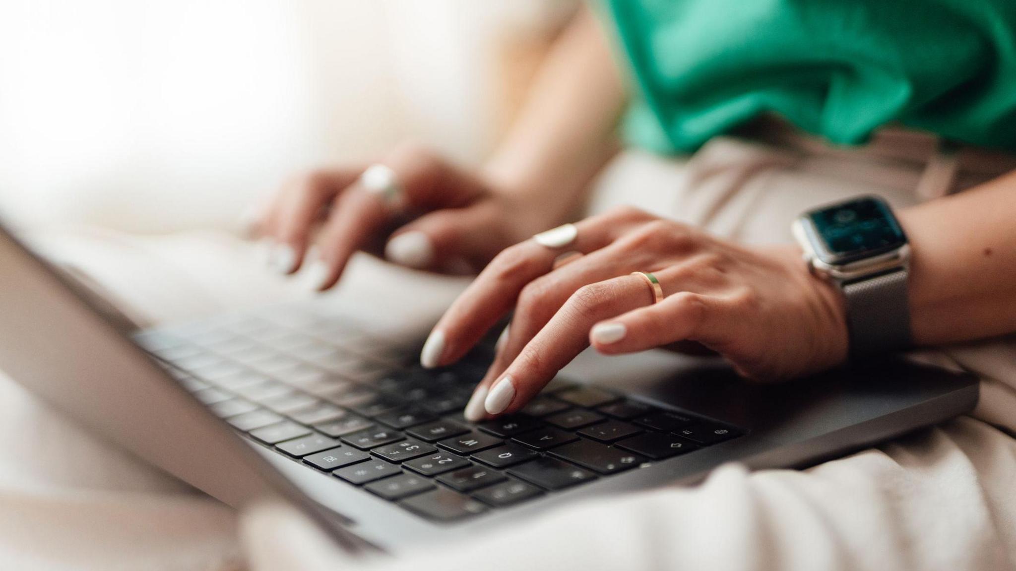 Someone's hands typing on a laptop keyboard. they have white nails and are wearing gold jewellery. They have an apple watch on their rest and are wearing a green top and beige trousers. The laptop is silver.