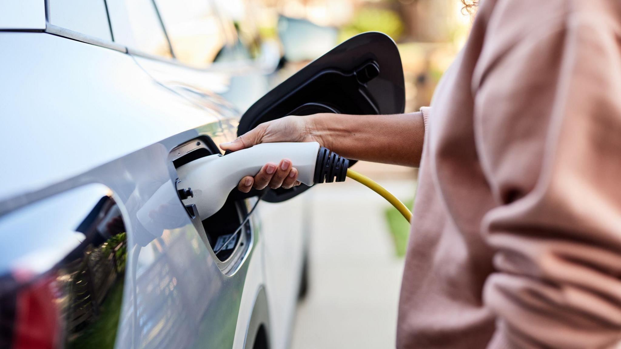 A woman in a pink jumper charges a white car. It is a close-up photo and only the woman's torso and arm can be seen.