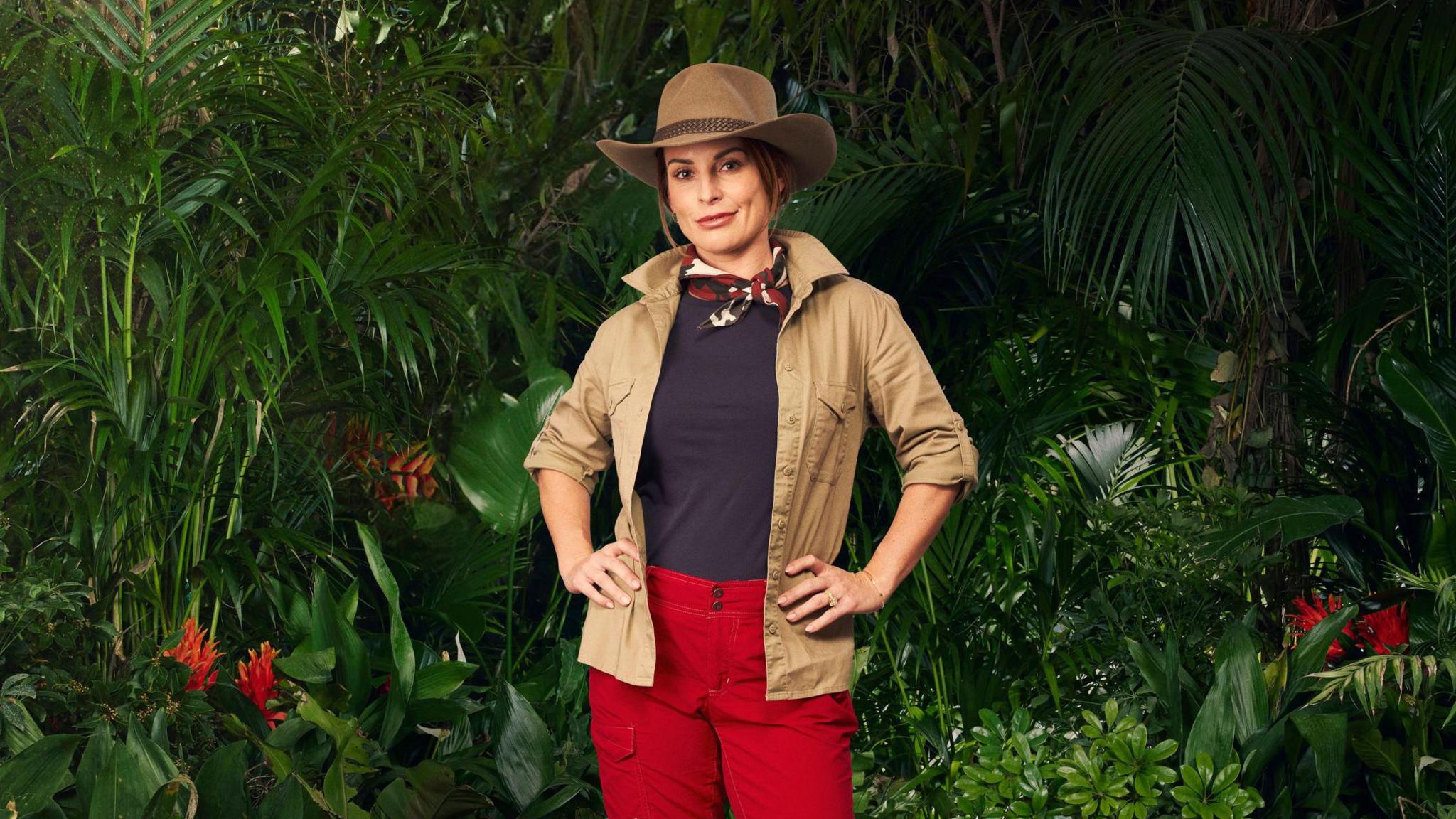 Coleen Rooney wearing her jungle outfit of red trousers and earth-toned jacket and hat, in a jungle scene surrounded by plants