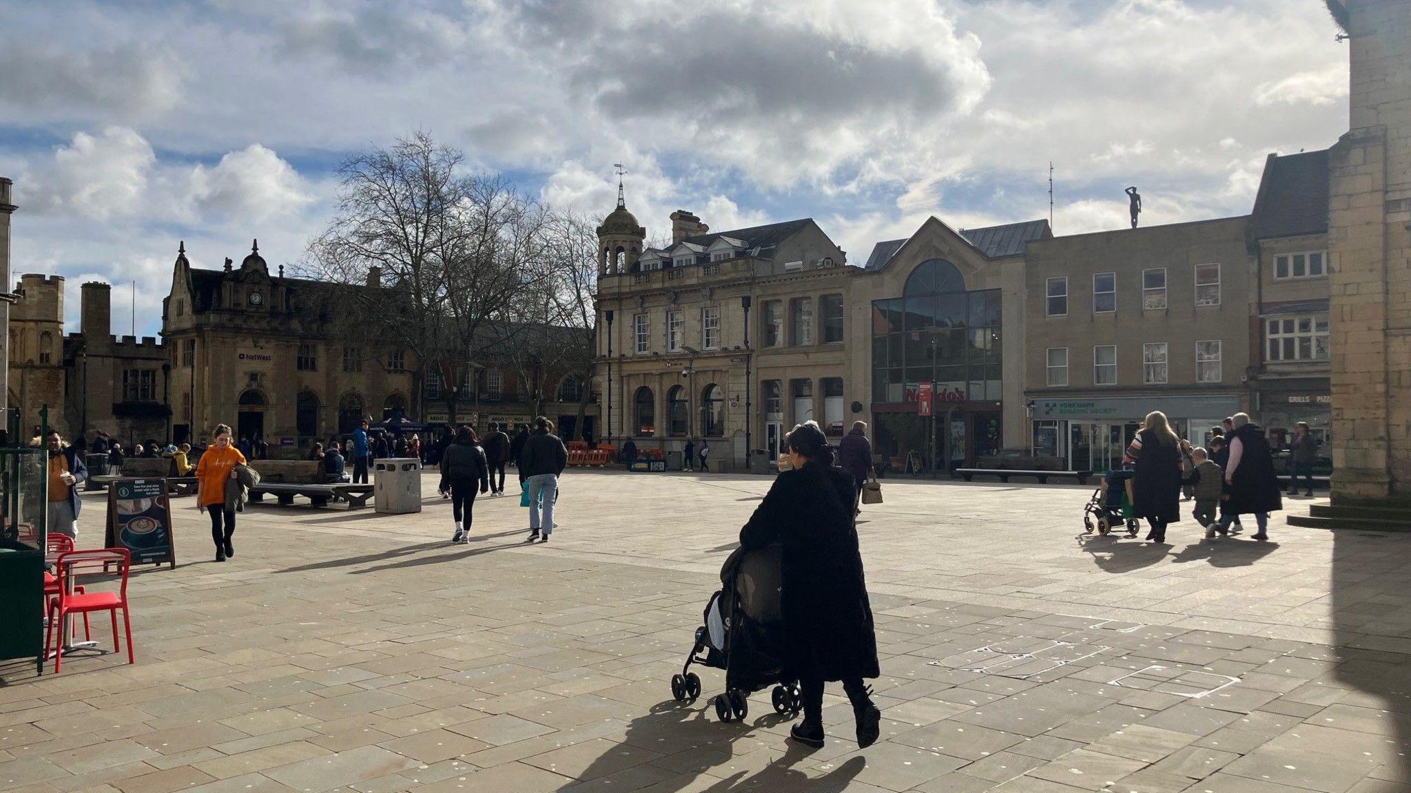 Peterborough's Cathedral Square with people walking around the square under a clouded sky - lined with buildings in the background.