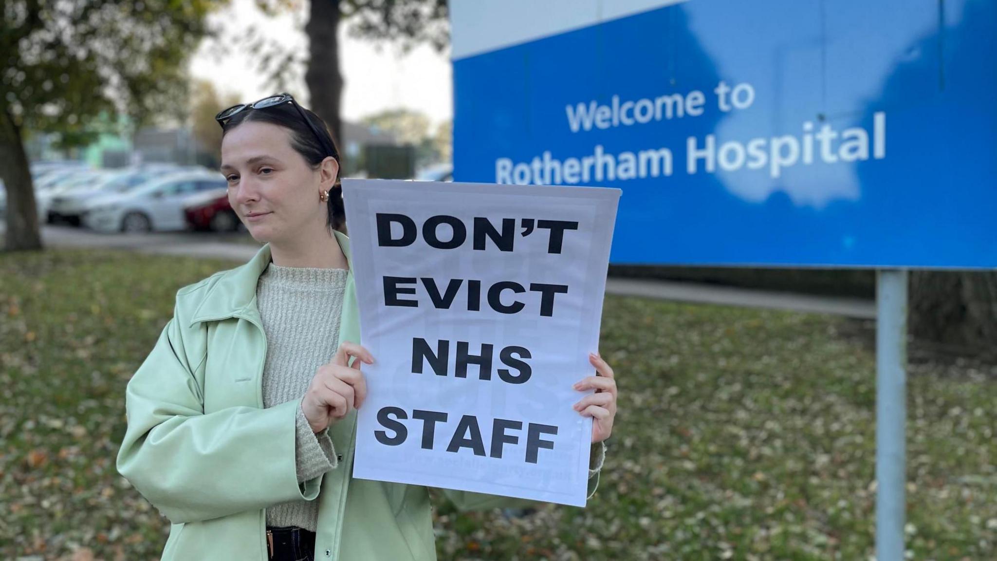 A young woman stands in front of a hospital holding a sign that reads: "Don't evict NHS staff." The woman is wearing a light green coat and a cream-coloured jumper, with sunglasses perched on top of her head.