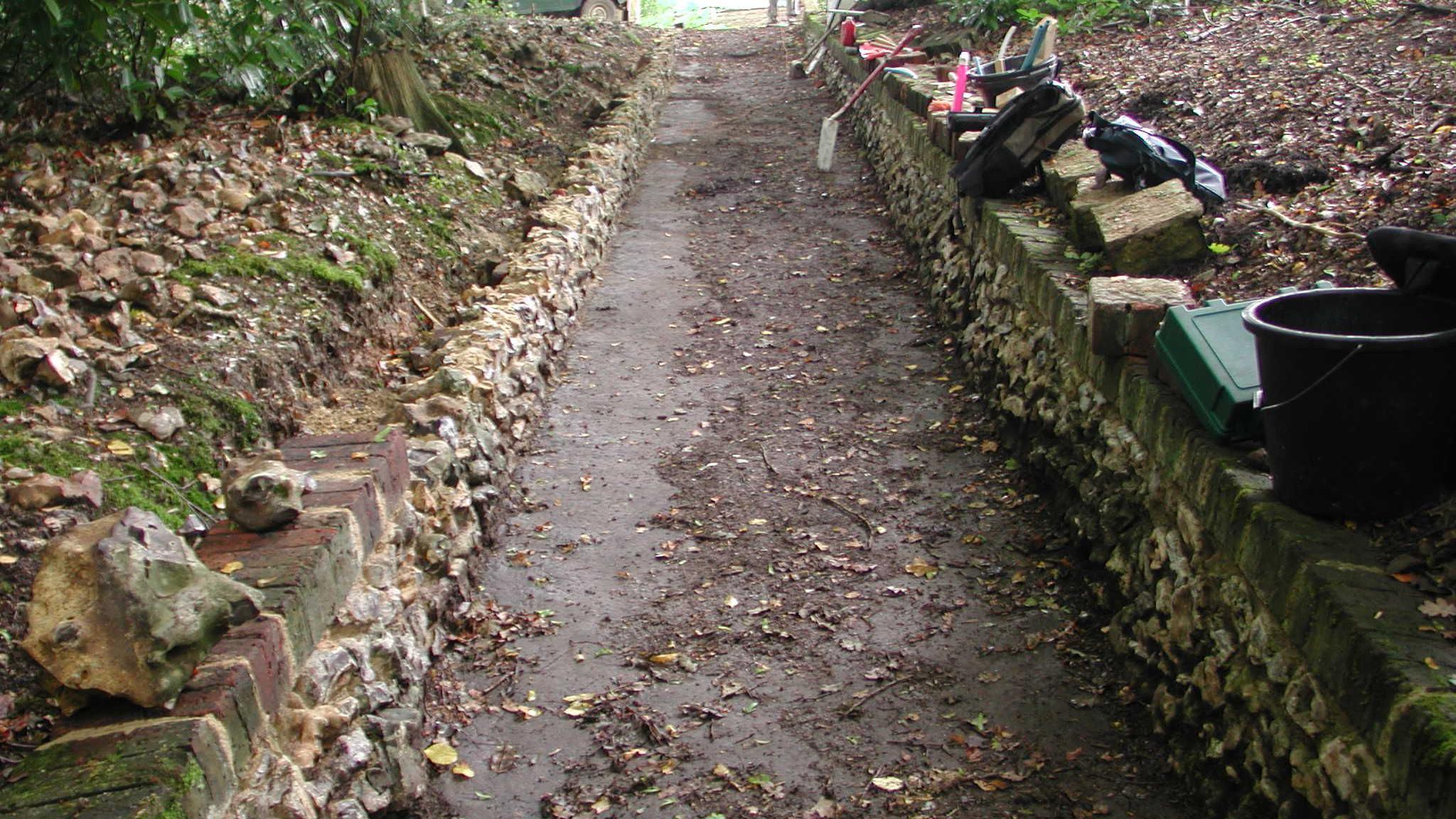 Flint walls on a carriage ride, that have been restored, showing materials left to the right, a pathway, with mud on it, and trees, leaves and foliage on either side. 