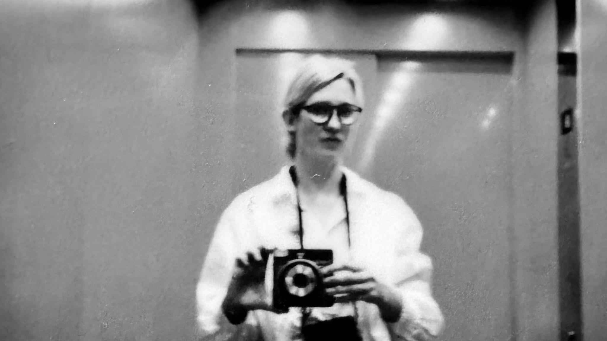 Black and white polaroid style headshot of Gemma Briggs. She is using a camera to take a photo of herself in a mirror. She has light hair, glasses and wears a white blouse. 