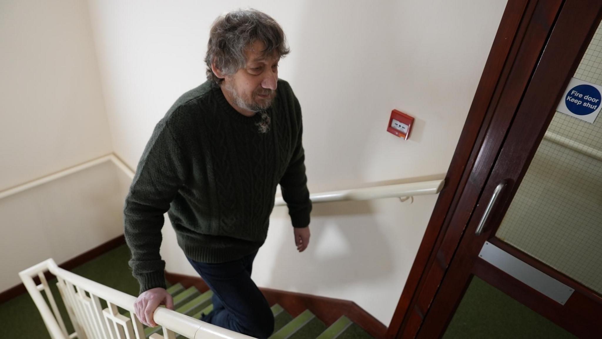 Daniel Evans-Smith walking up the stairs to his flat in Northampton. He is holding onto a cream banister and is wearing a green wool jumper and navy cordroy trousers. His beard his slightly plaited and his hair is greying.