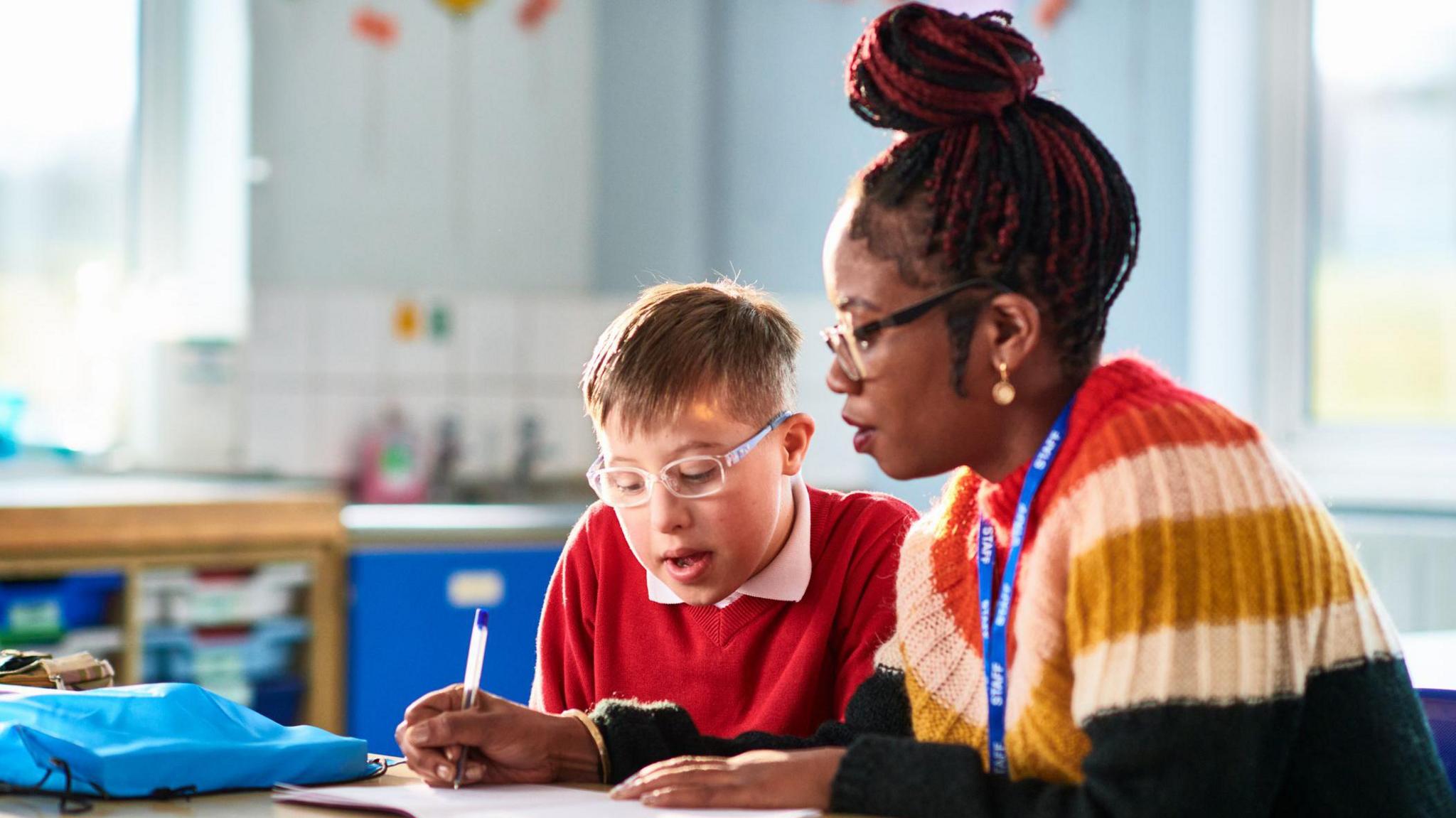 Female support worker assisting child with special educational needs, writing in his book as he watches and concentrates