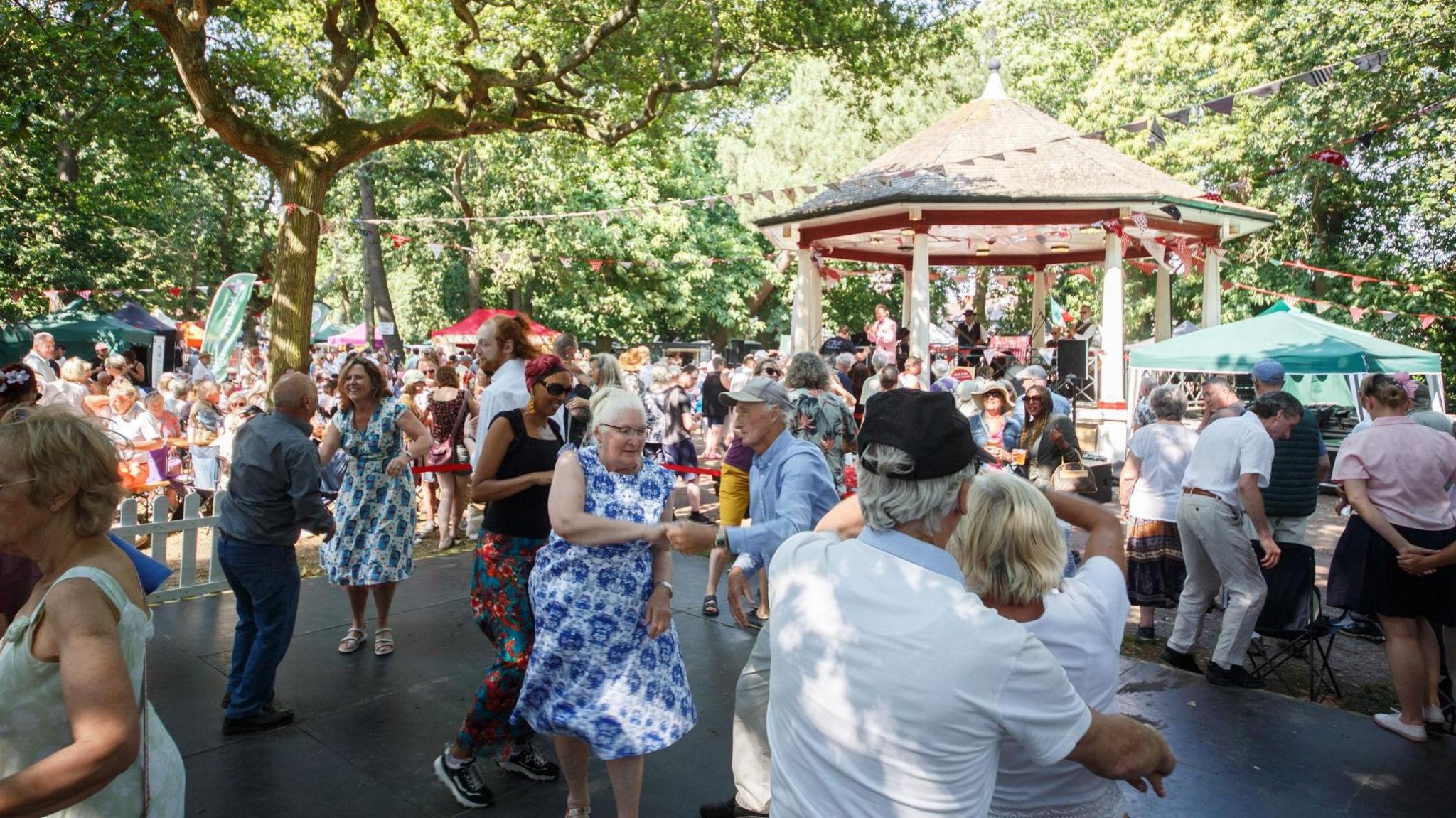 Crowds of people at the festival in 2024. In the foreground is a dancefloor area with couples jive dancing. In the background is a band playing in a bandstand.