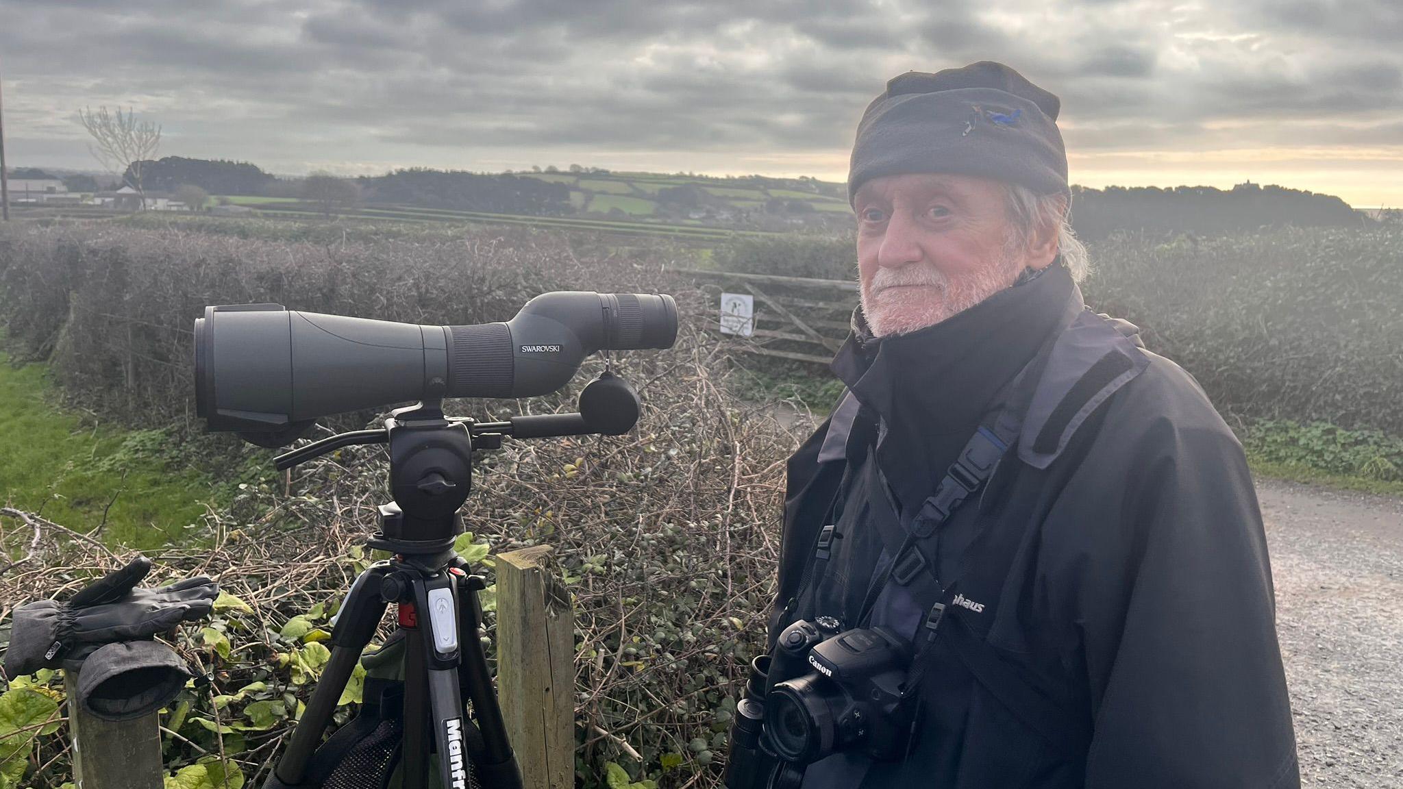 Man with short white beard in warm hat standing beside a telescope beside a field.