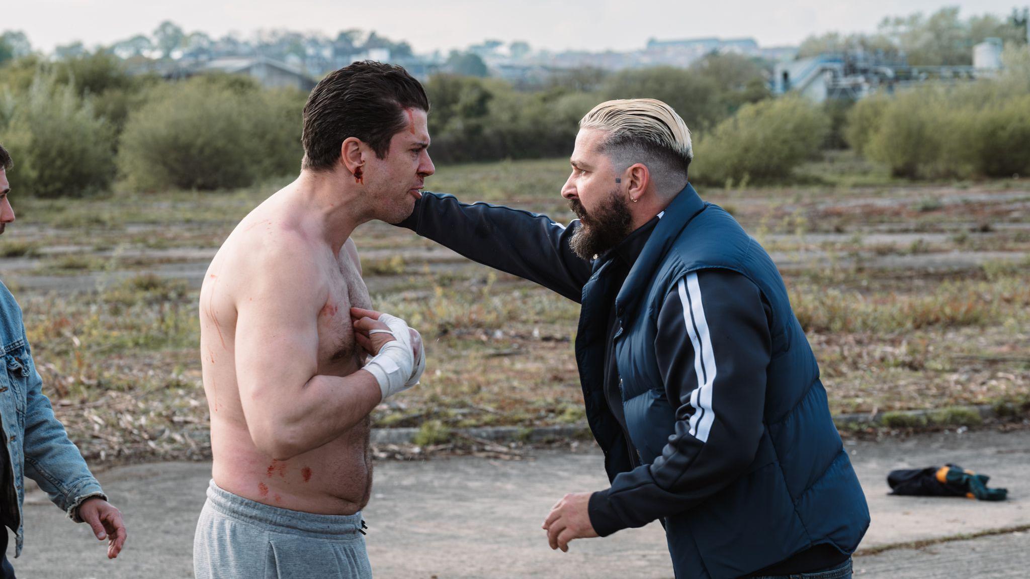 Shia LaBeouf and Toby Kebell stand angrily looking at eachother face to face on a yard. Labeouf stands on the right hand side with his hand on Kebell's shoulder. has bleached blonde hair slicked back and cut short on the sides. He wears an earring and has a dark brown long beard. He wears a black zip up top with two white stripes on the sleeves and a black gilet. Kebell is topless with grey joggers on, he has blood coming from his ear and his hands are bandaged. 