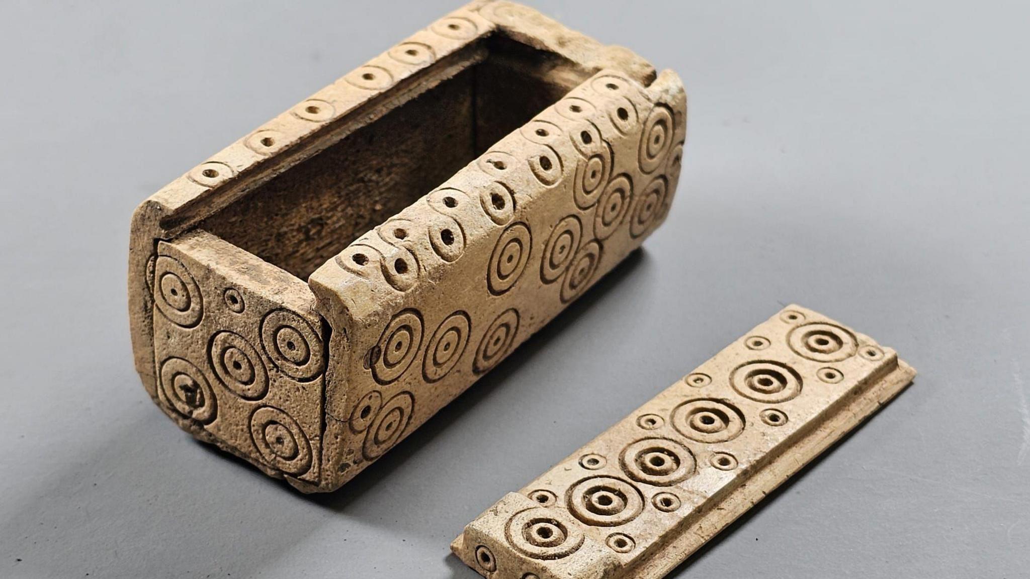 A small box made out of bone with round circles carved into the side for decoration. It is next to a sliding lid that closes the box.