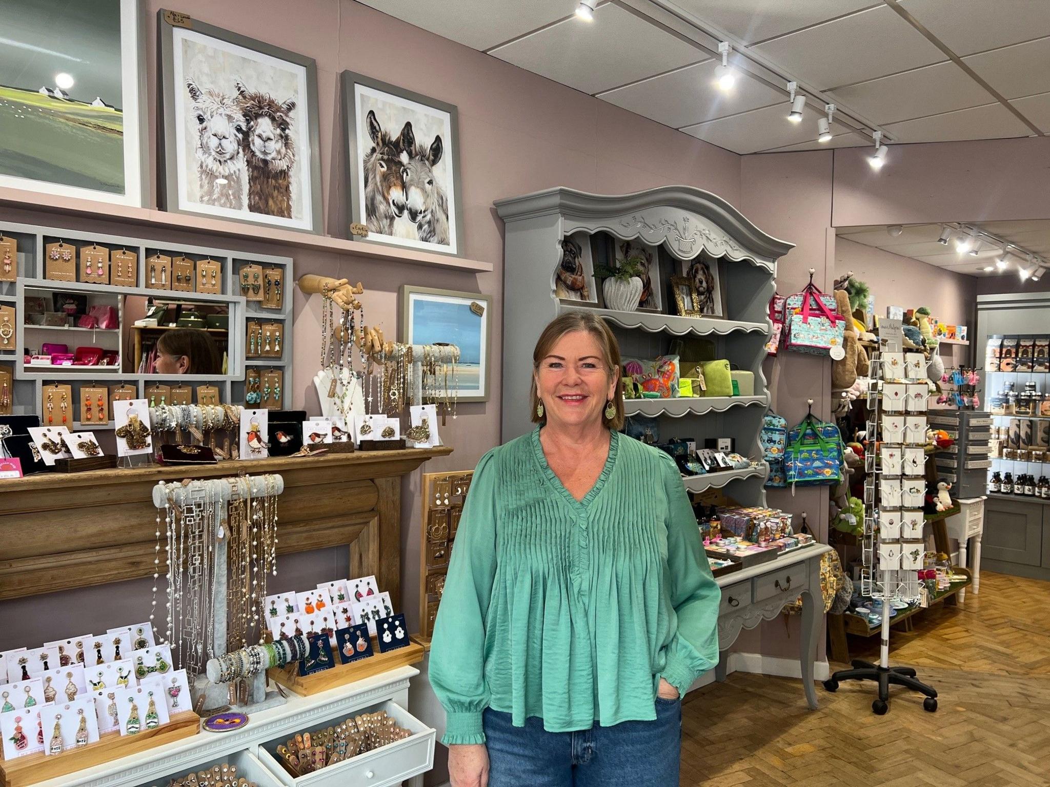 A woman with dark hair wearing a green top is smiling at the camera. Many gift items are behind her next to a wall.