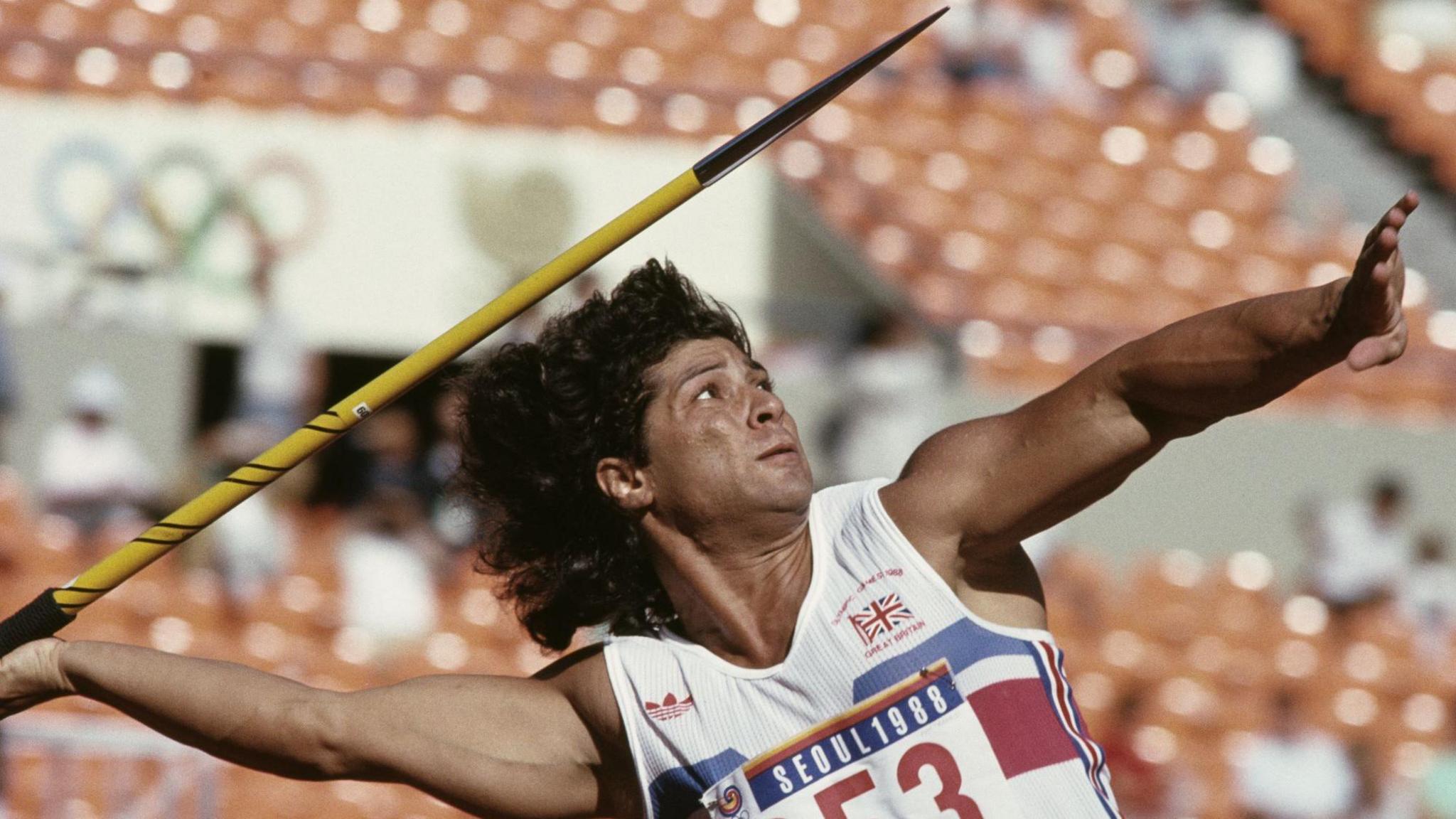 Fatima Whitbread poised with a javelin which she is about to throw. She is in a stadium. 