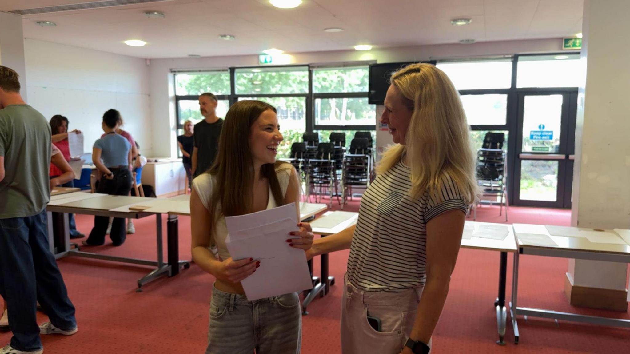 A girl with brown hair holding a piece of paper and smiling. She is looking at the woman to the right, who is blonde, and has her arm against her arm. There are tables behind them with pieces of paper on them. There are other people around them opening A level results. The floor is red carpet.