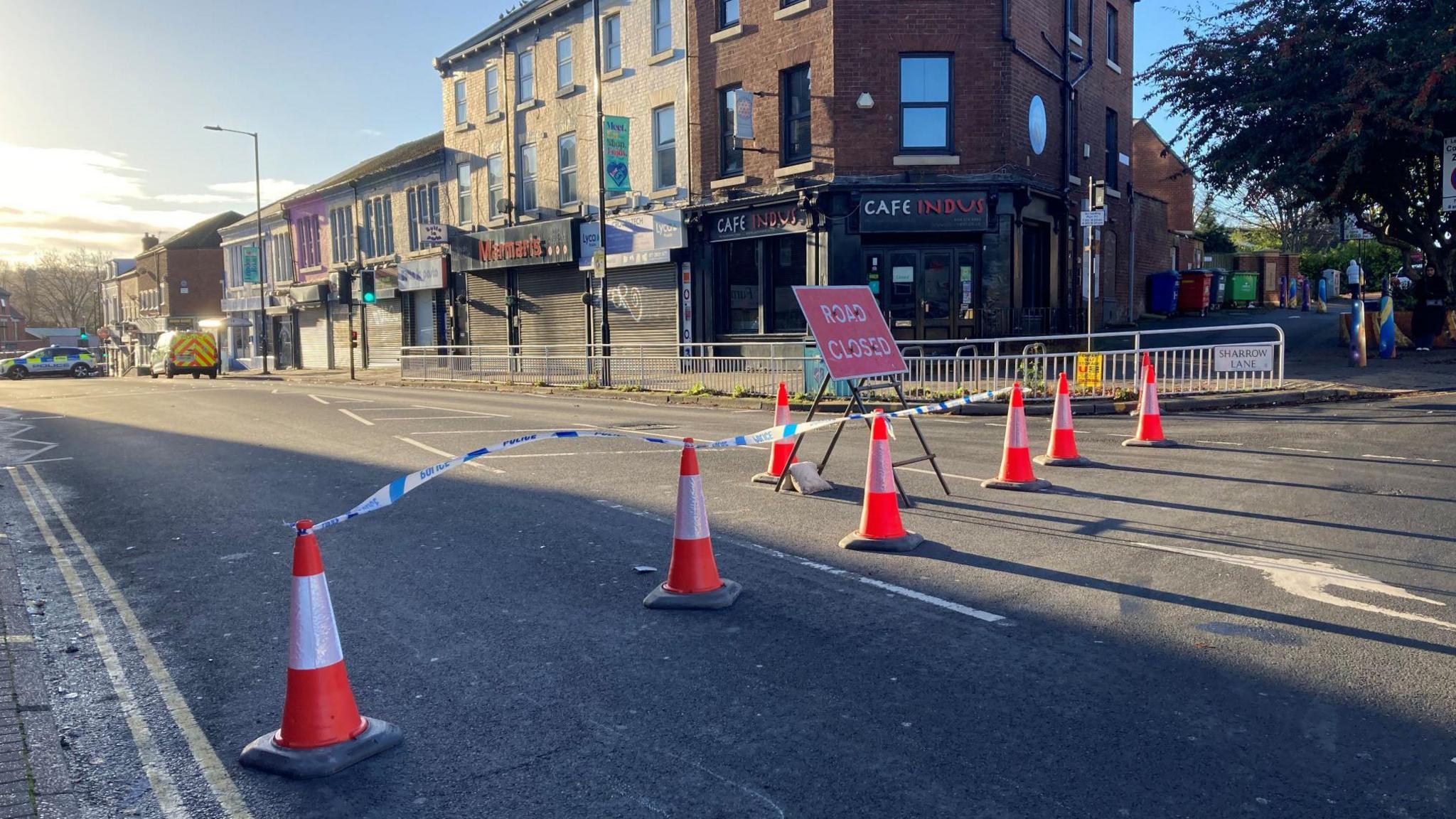 A street cordoned off with blue and white police tape and orange and white cones. There is a sign in the middle of road which reads 'road closed'. In the background are two parked police vehicles.