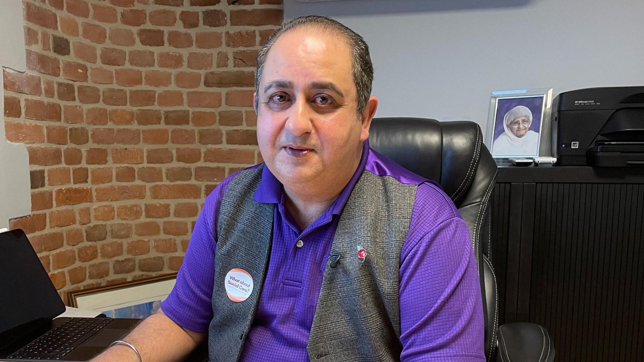 Raj Sehgal in a purple polo shirt and grey and purple waistcoat. He is sitting at a desk with a brick wall behind him.