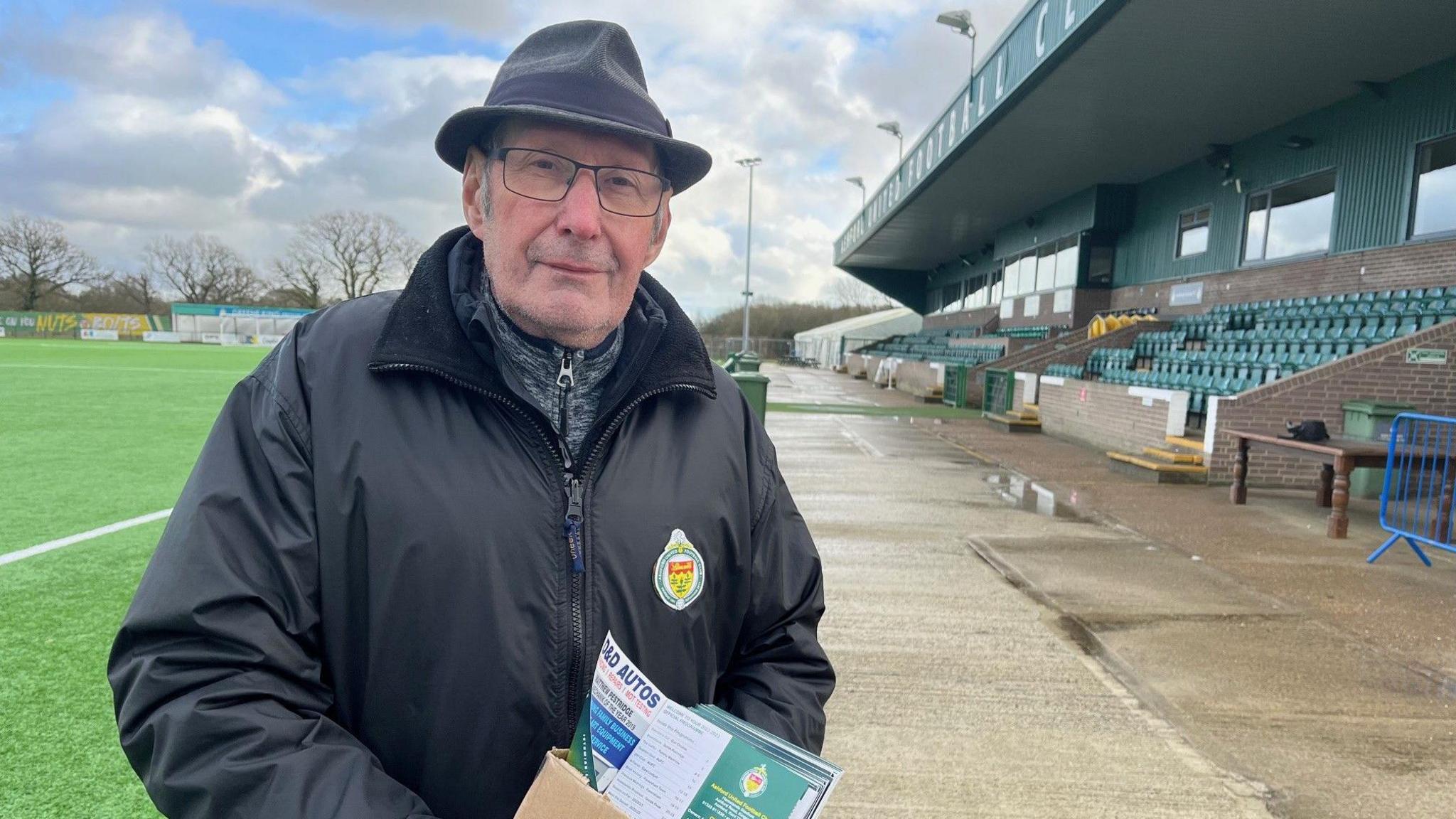 A man wearing a black coat and hat in a football stadium holding a cardboard box of football programmes.