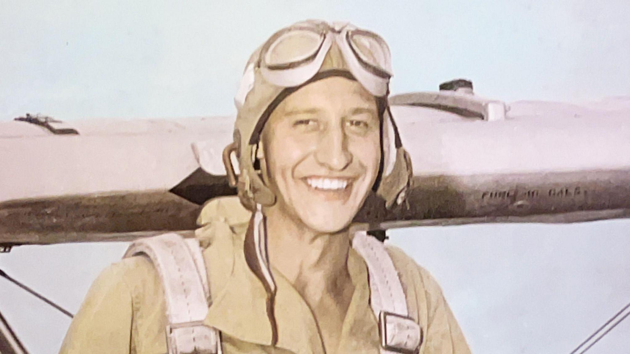 Raymond King is wearing a beige flight suit, flying goggles and a flying cap. He is smiling ear to ear and standing in front of the wing of a plane