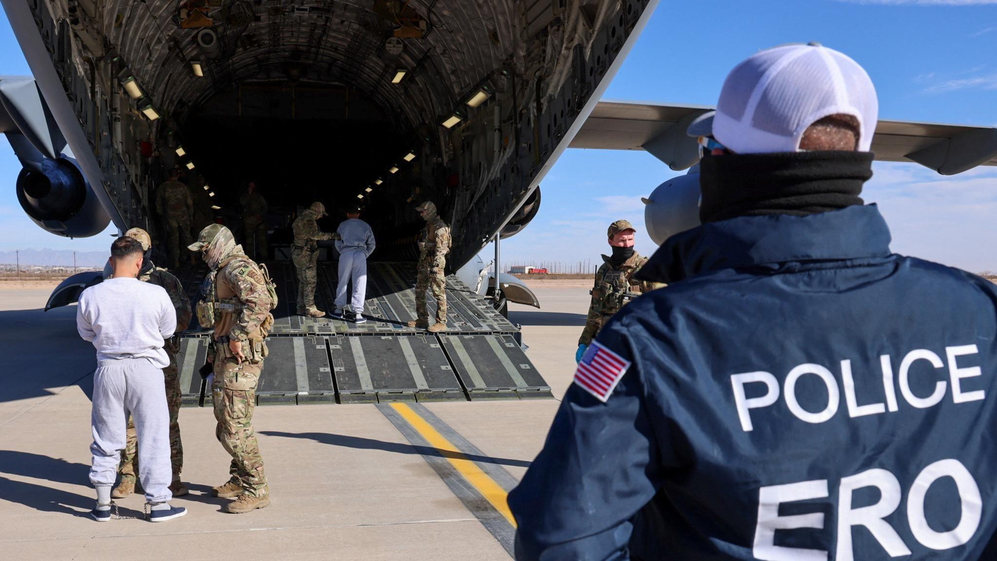 A police officer looks on as two men in grey tracksuits are taken on board a US military aircraft by officers in camouflage uniforms