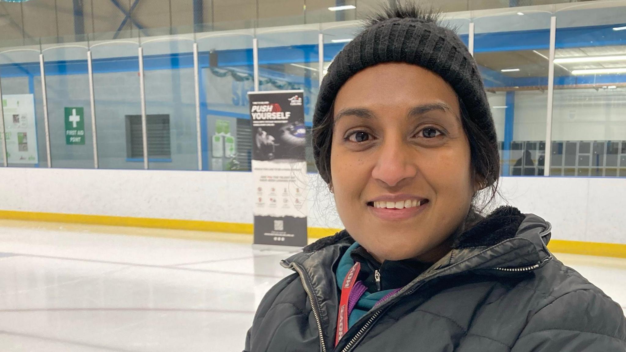 Tania Nadarajah is on an ice rink, smiling at the camera. She is wearing a dark-coloured jacket and a bobble hat