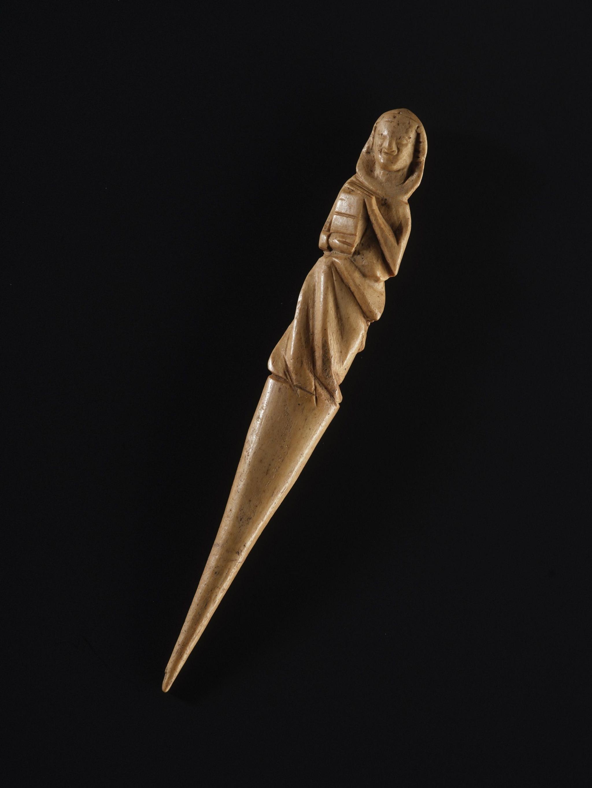 A close up of the gravoir, a needle-shaped ornately carved piece of red deer antler. The carving is of what looks like a hooded figure holding a book. The object is photographed against a black background.