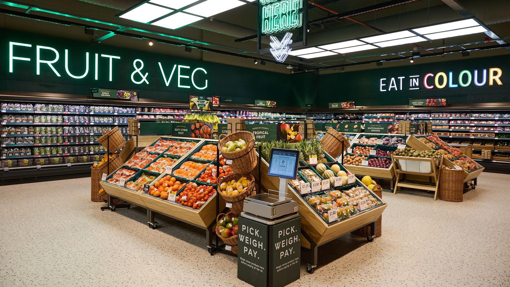 The fruit and vegetable section of a Marks and Spencer store. There are two islands with brightly coloured displays of fruit and vegetables, and a set of self-service weighing scales in the foreground.
