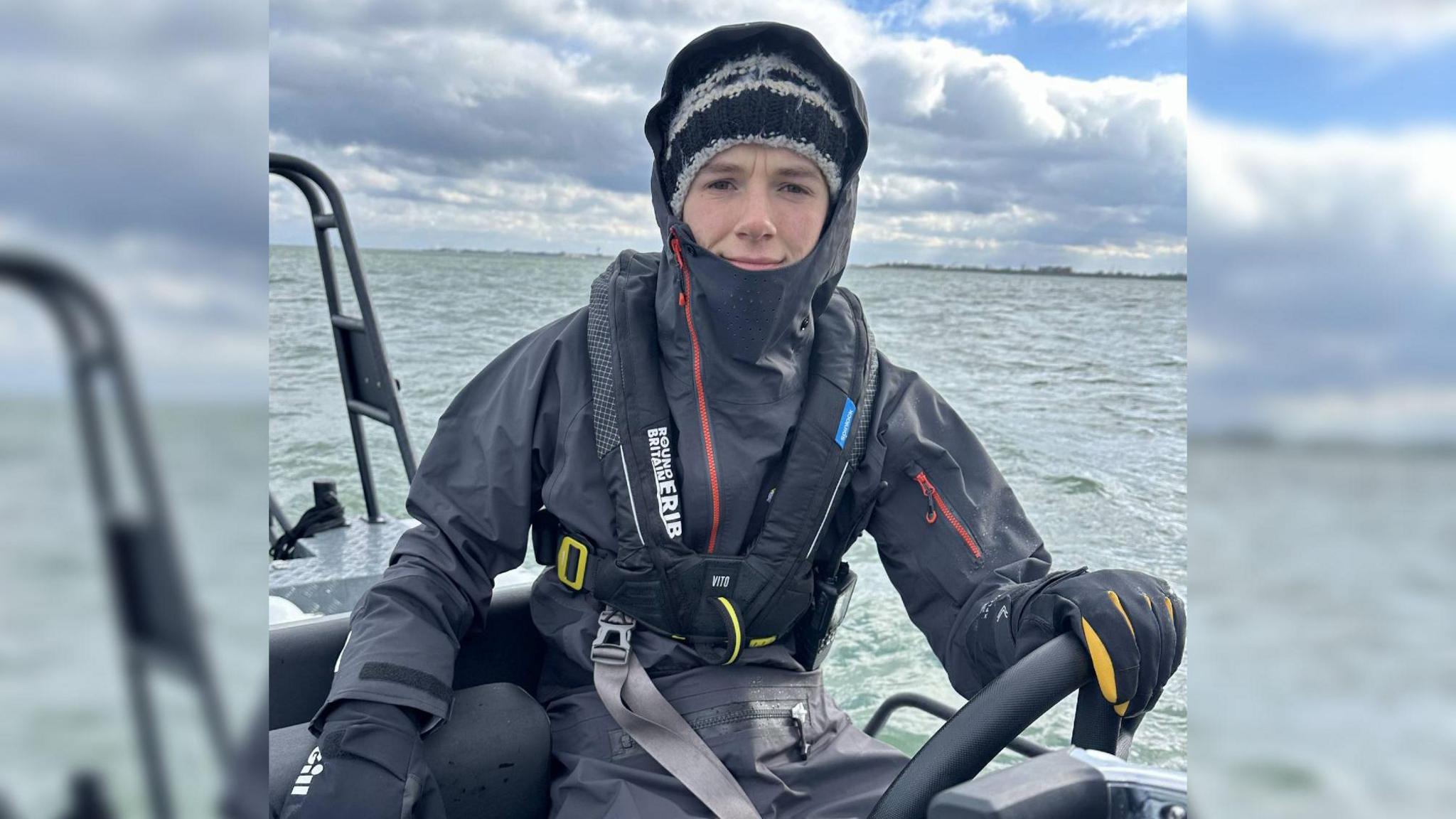 Harry Besley sitting behind the wheel of an electric RIB boat out at sea. He is wearing waterproof gloves and overalls with his hood up