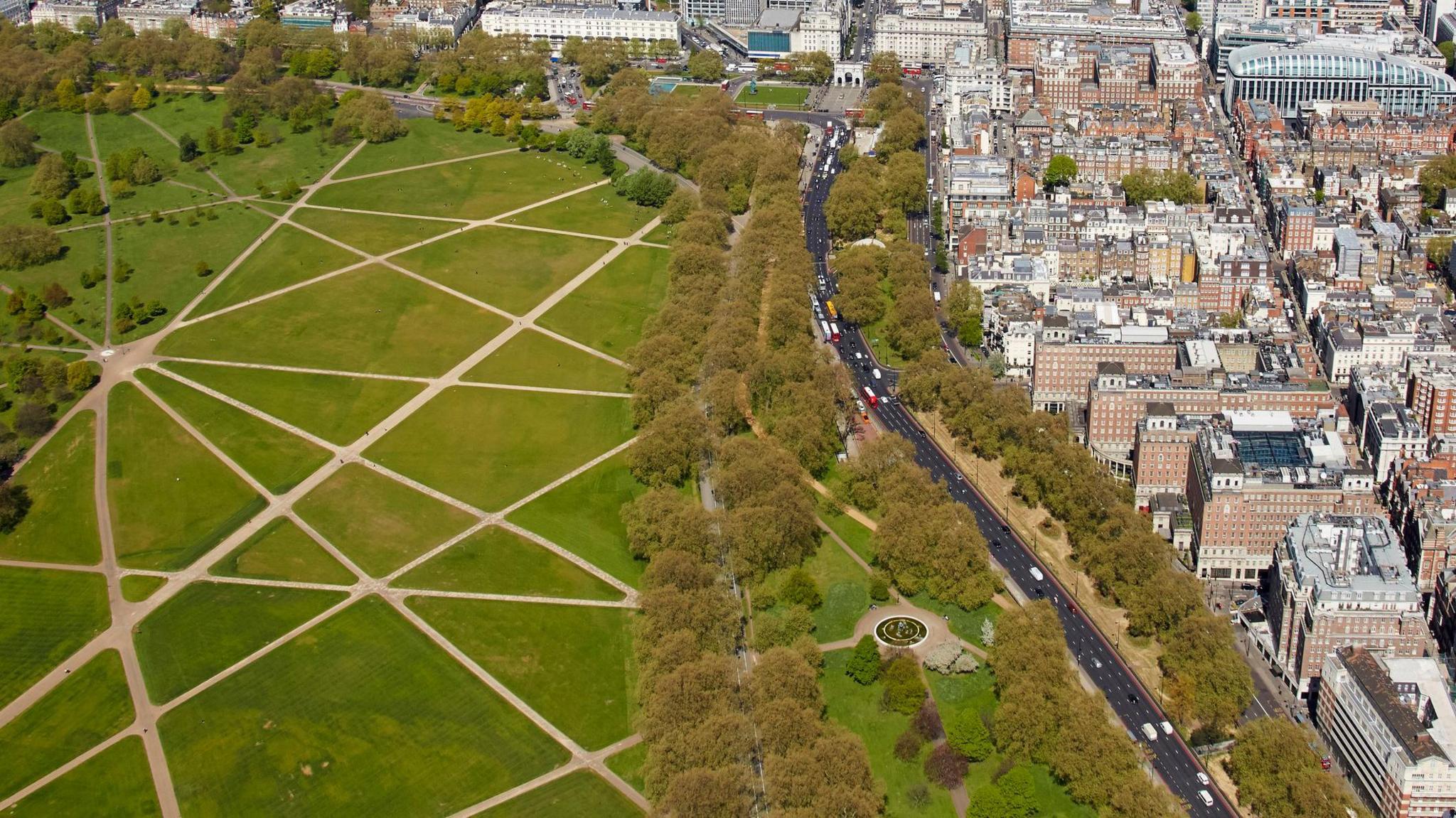 Aerial view of Park Lane in London, with Hyde Park on the left featuring criss-crossing footpaths over open green space. A tree-lined road separates the park from dense urban buildings and streets on the right. Traffic, including red buses, is visible along Park Lane