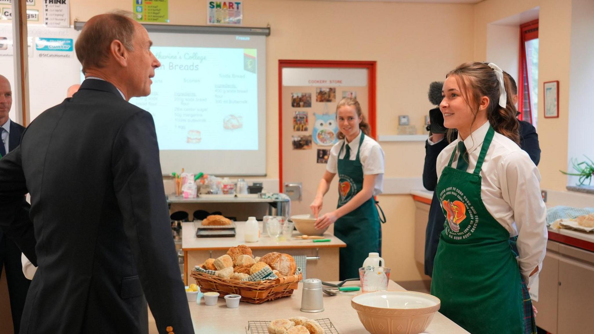 The Duke of Edinburgh, Prince Edward, in a classroom, chatting to a girl in a green apron. There are mixing bowls and loaves of bread on the bench. 